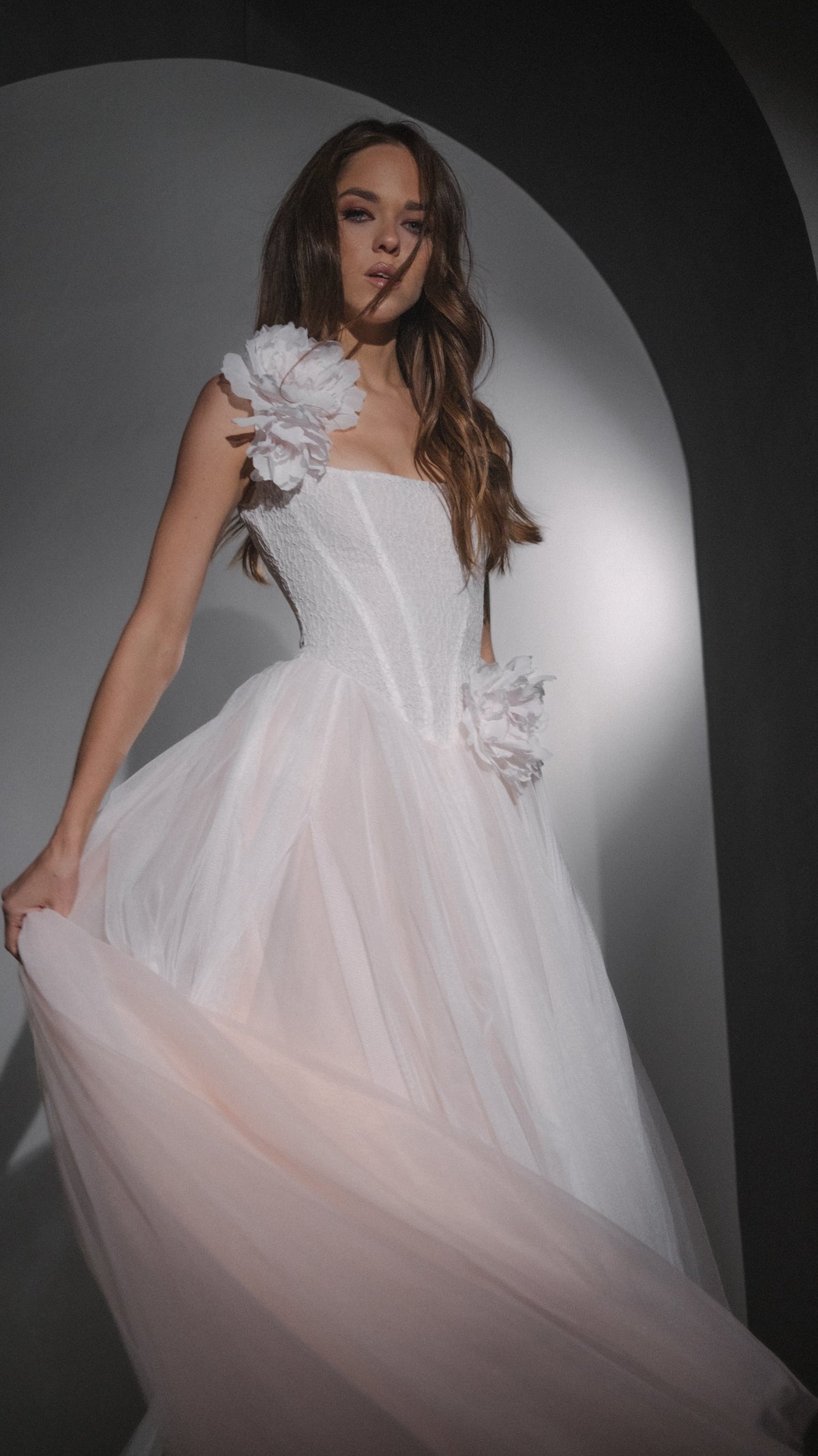 Model swirling the skirt of a blush basque waist wedding dress, silk flowers on the strap and hip, studio arch, Chrysanthemum