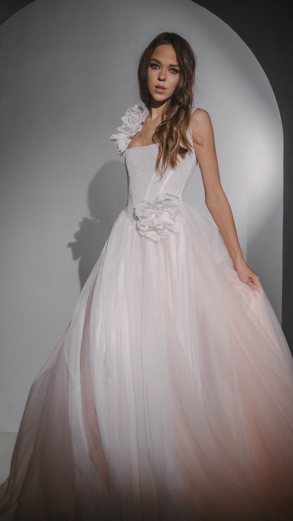 Front view of a blush wedding dress with dropped corset bodice and a silk flower at the hip, model in a pale arch, Chrysanthemum