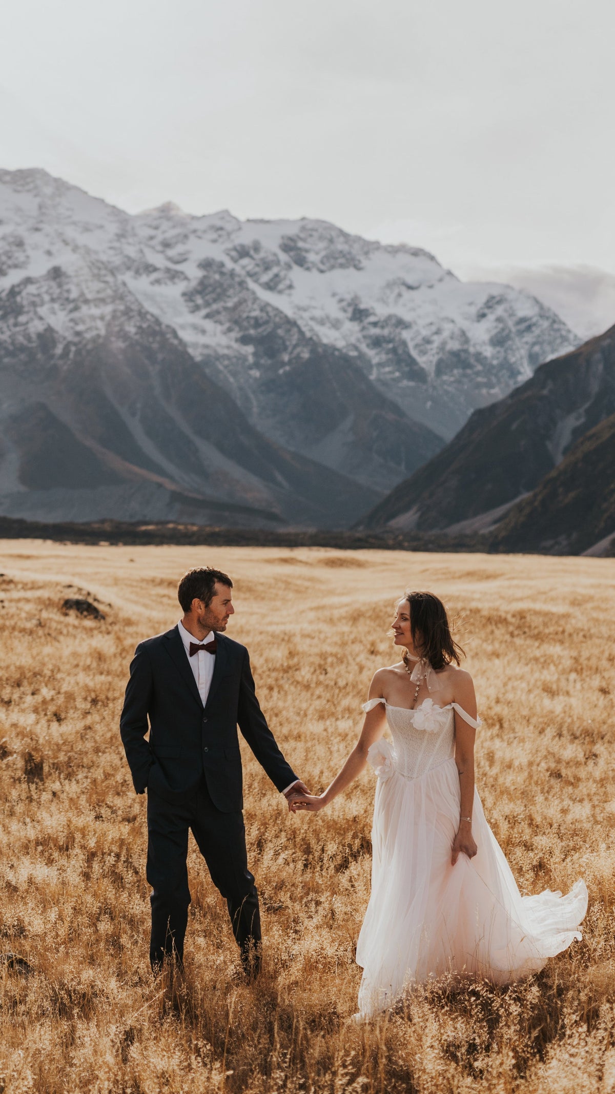 Bride and groom holding hands in a golden valley under snowy peaks, off-shoulder blush corset wedding dress, Chrysanthemum