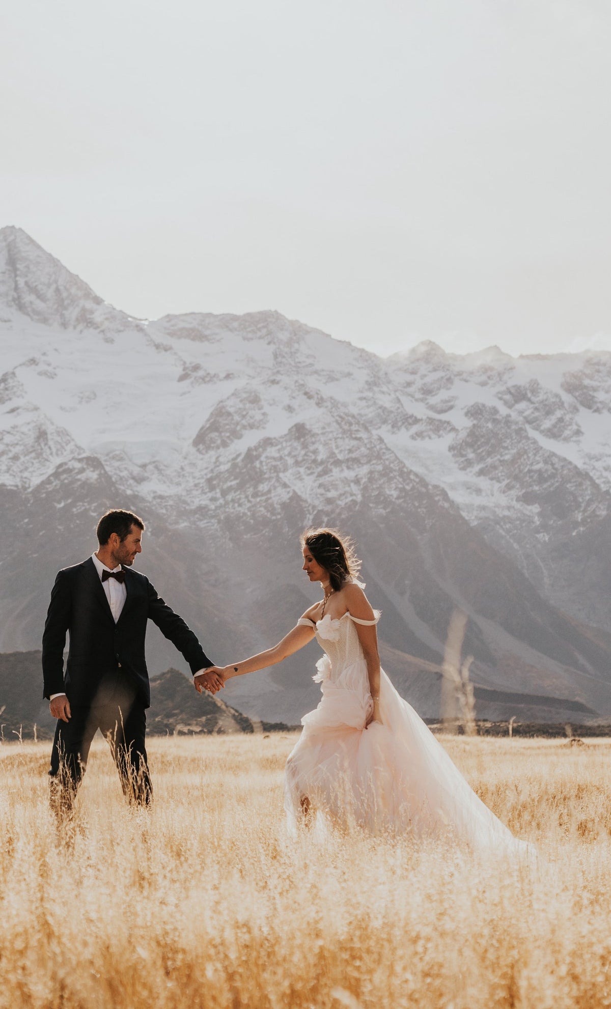 Bride leading the groom through tall grass below snowy mountains, blush tulle wedding dress skirt in motion, Chrysanthemum