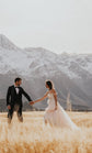 Bride leading the groom through tall grass below snowy mountains, blush tulle wedding dress skirt in motion, Chrysanthemum