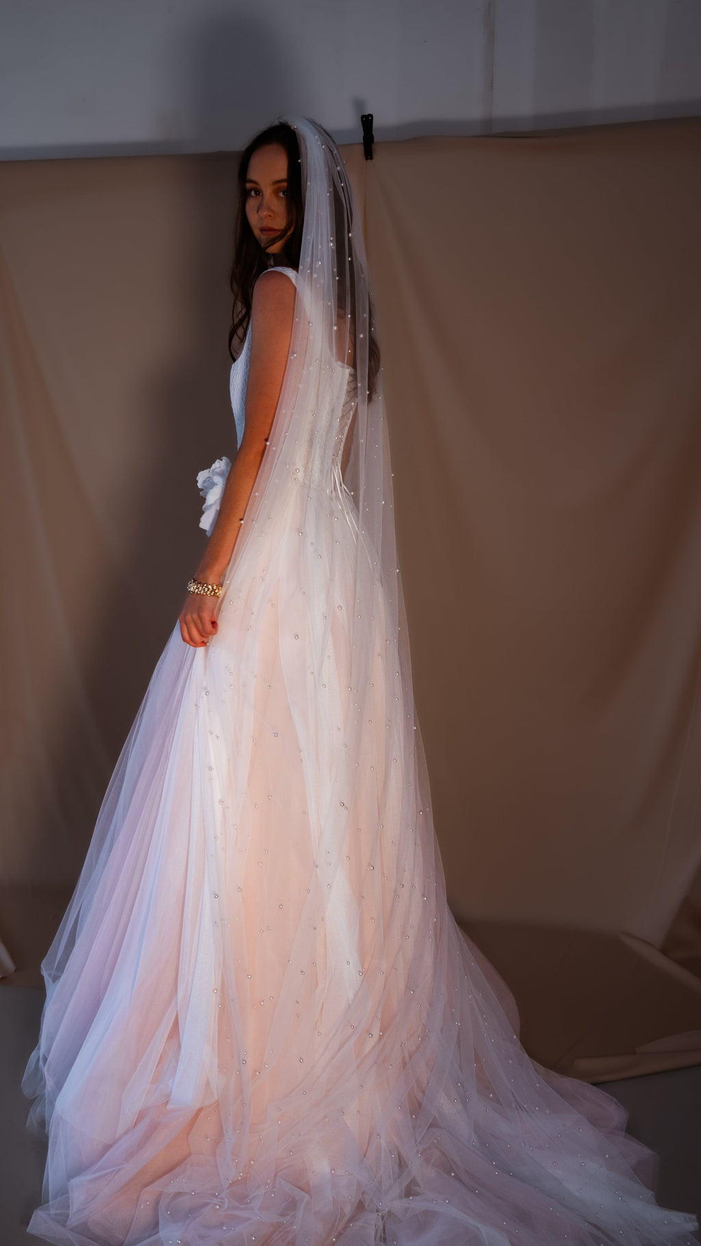 Bride looking over her shoulder in a blush corset wedding dress, long pearl-dotted veil over the tulle skirt, Chrysanthemum