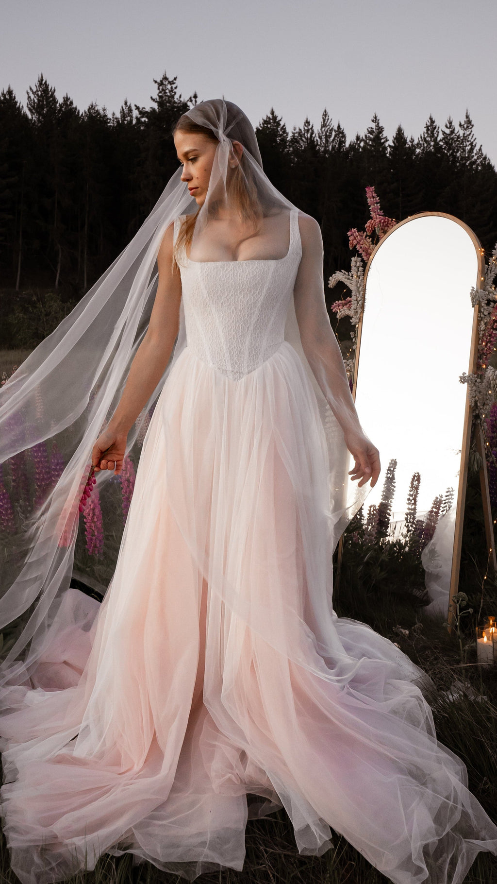 Bride under a long sheer veil in a blush corset wedding dress with a mirror and lupines in a field at dusk, Chrysanthemum