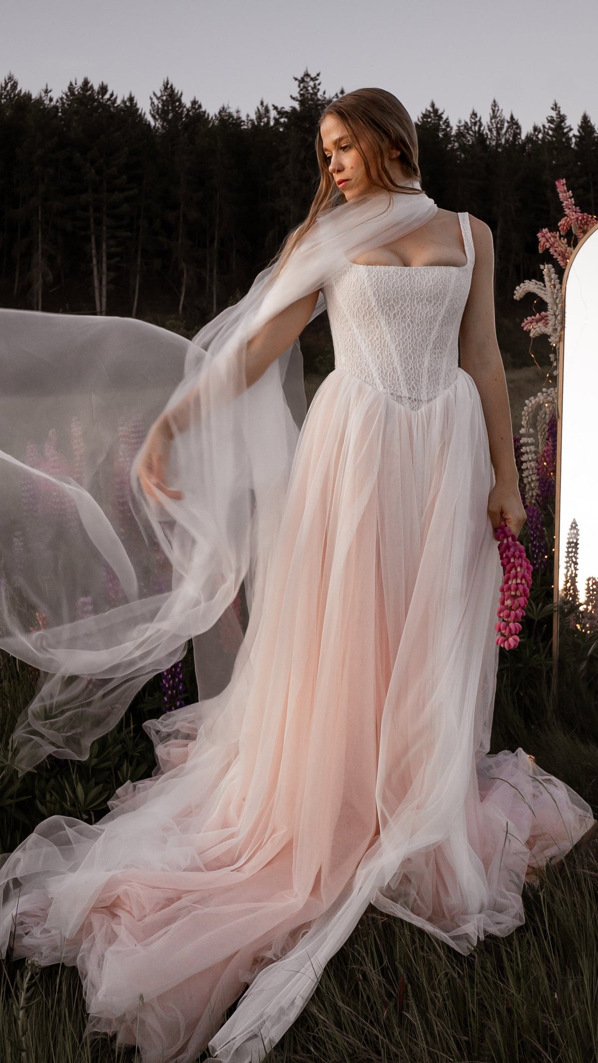 Bride holding a pink lupine with a long tulle veil draped over one shoulder, blush corset wedding dress, Chrysanthemum