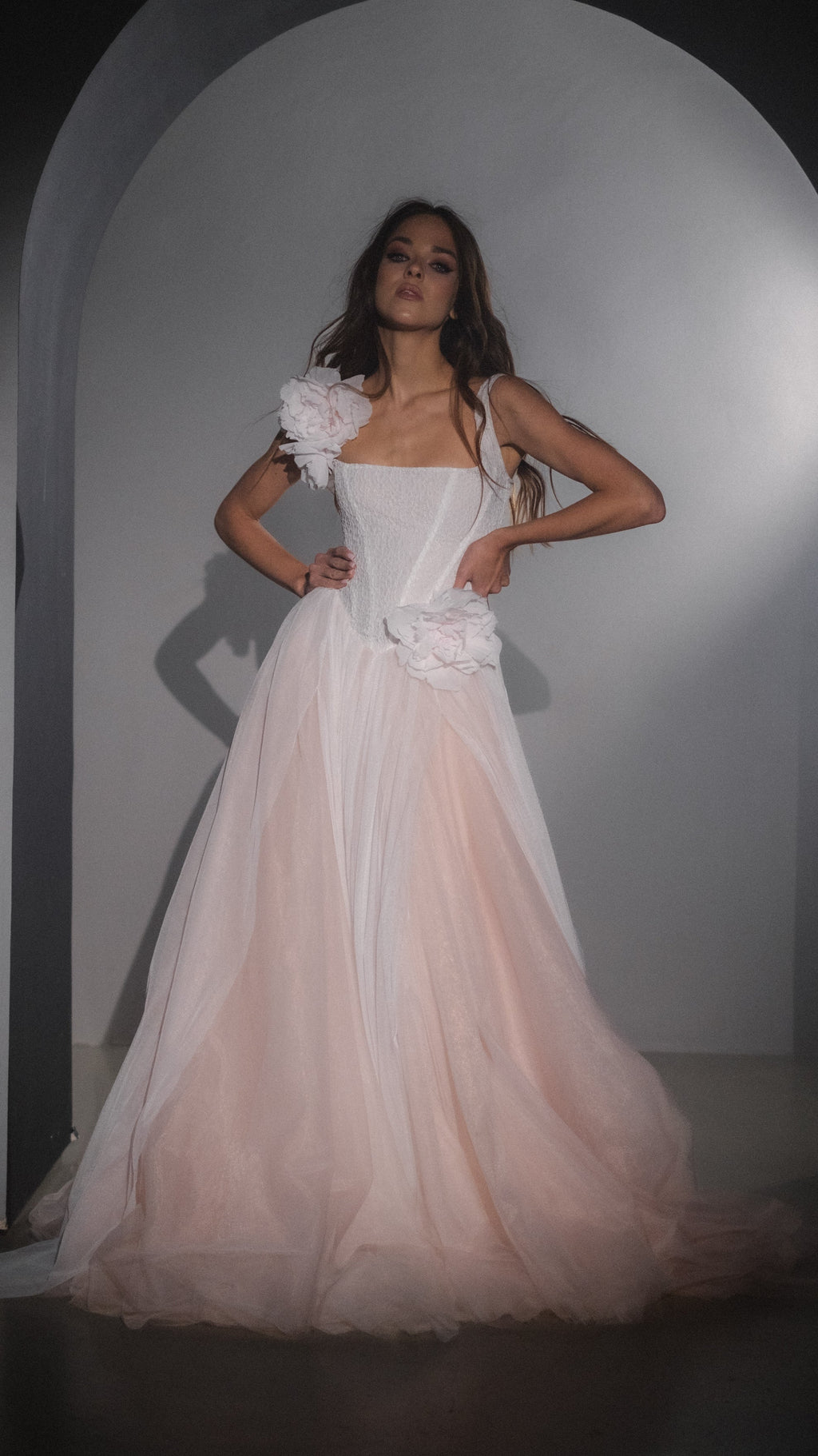 Model with hands on hips in an arched alcove, blush pink corset wedding dress with removable silk flowers and tulle skirt, Chrysanthemum