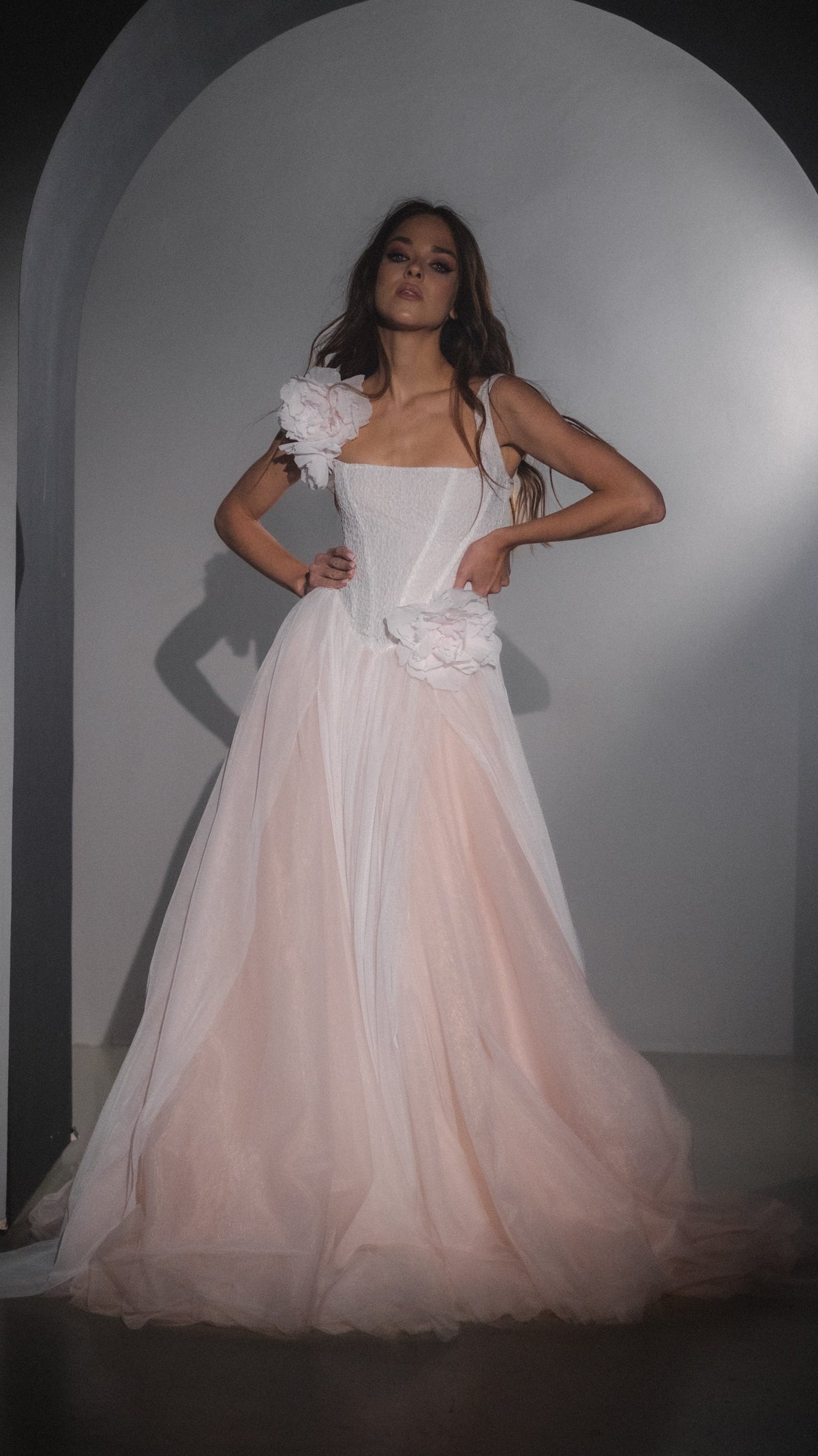 Model with hands on hips in an arched alcove, blush pink corset wedding dress with removable silk flowers and tulle skirt, Chrysanthemum