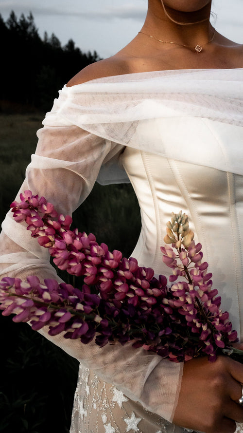 Close-up of off-shoulder detachable tulle sleeves tucked under a boned satin corset, pink lupines held across the body, Cloud Sleeves