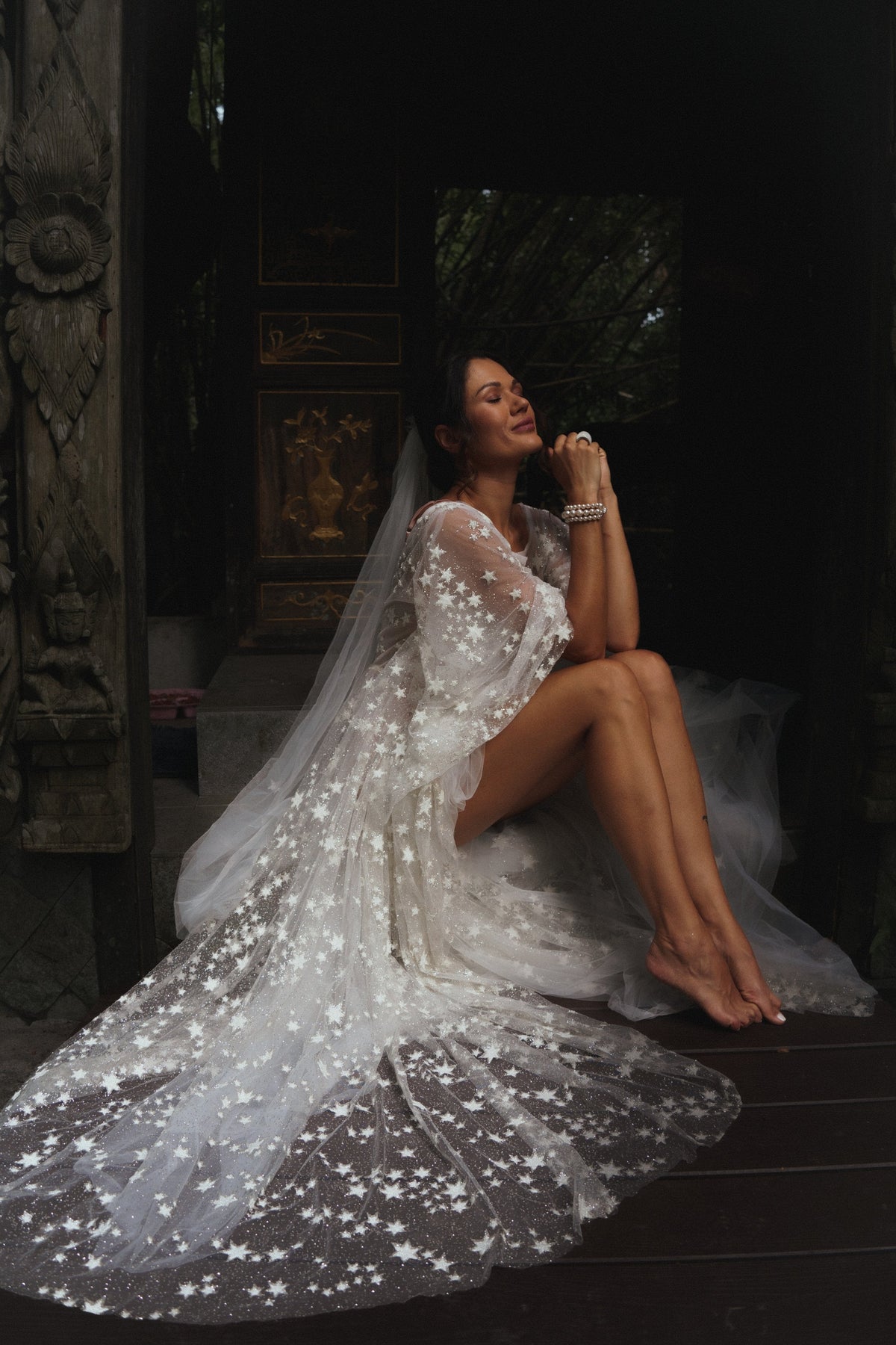 Bride seated barefoot in a dim courtyard, star-scattered cathedral wedding veil spread across the floor around her, Coastal Moonlight Veil