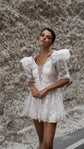 Front view of a short wedding dress with plunging neckline, star lace puff sleeves and a full skirt, rock wall behind, Cosmic Dancer