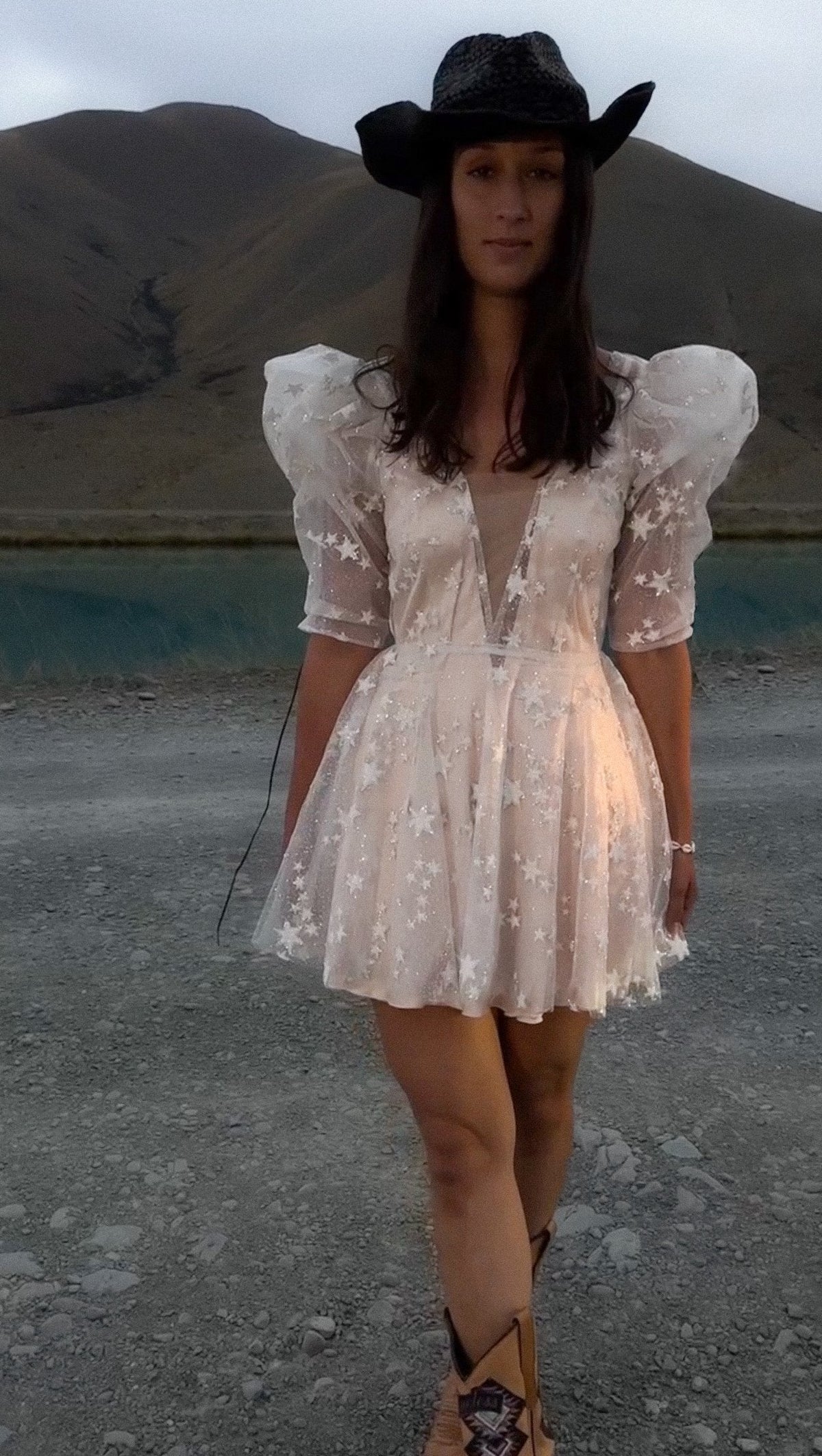 Bride in a black cowboy hat and boots wearing a star lace mini wedding dress with puff sleeves by a lake, Cosmic Dancer