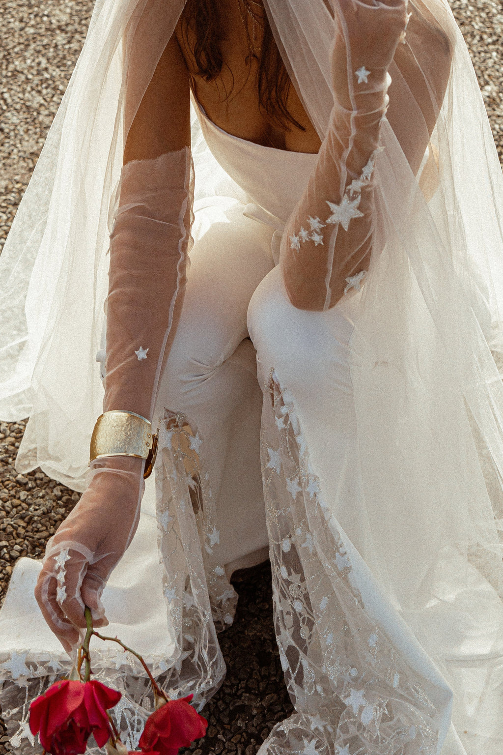 Sheer star tulle bridal gloves with a gold cuff, bride kneeling on gravel holding red roses under a long veil, Cosmic Gloves
