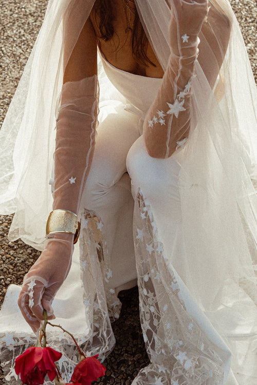 Sheer star tulle bridal gloves with a gold cuff, bride kneeling on gravel holding red roses under a long veil, Cosmic Gloves
