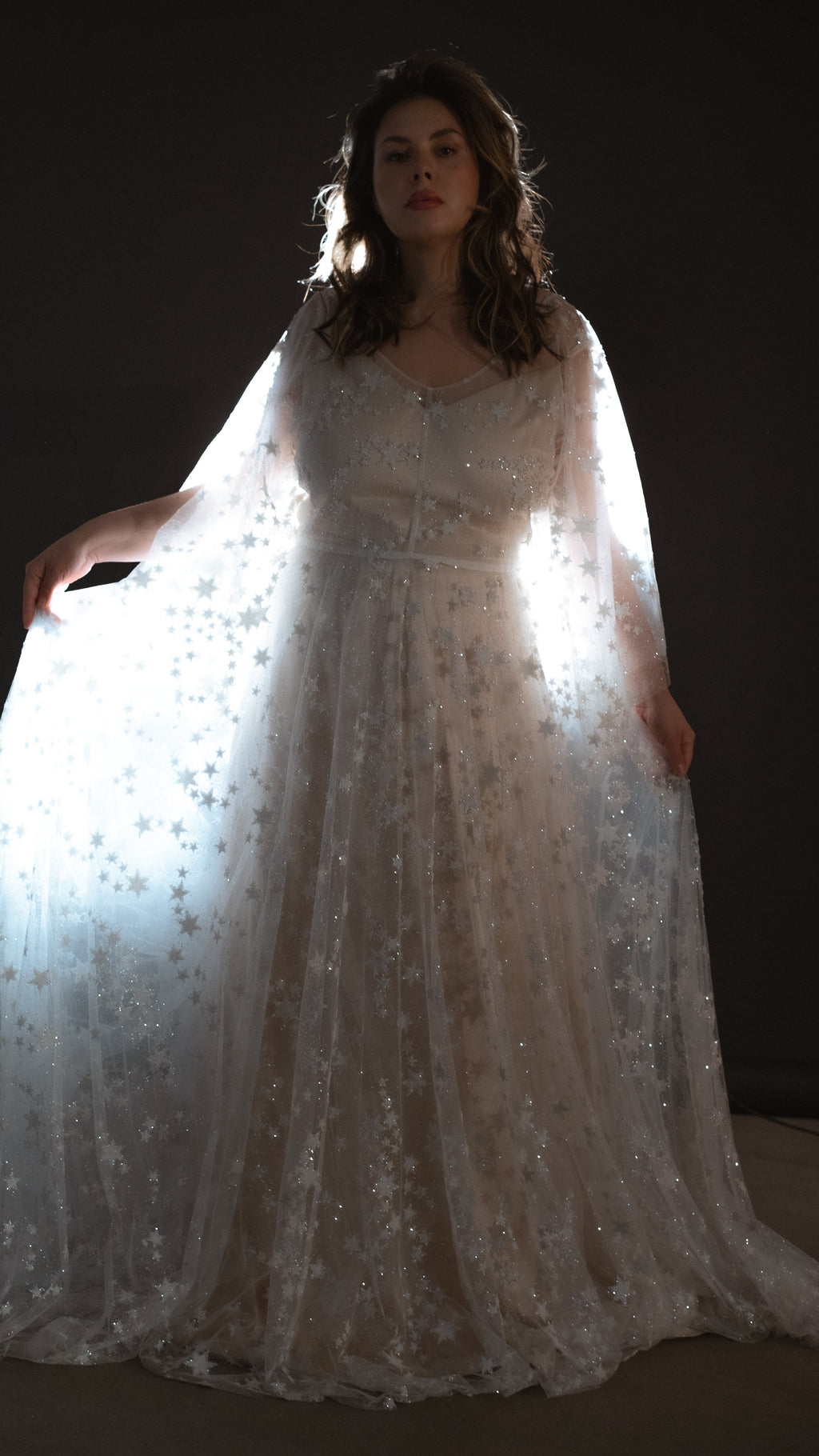 Glowing backlit star lace wedding dress with sheer bell sleeves held open against a black studio wall, Counting Stars 2.0