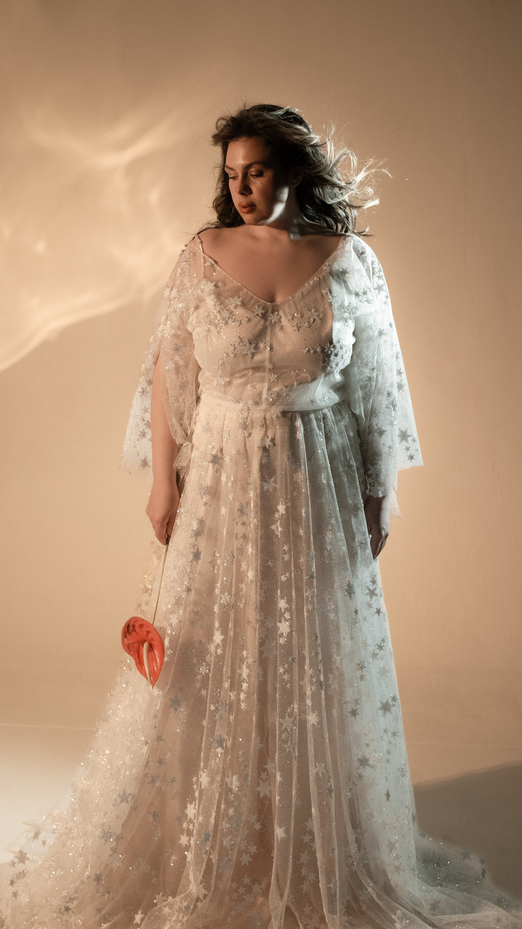 Bride looking down with a red anthurium at her side, star lace wedding dress with a scoop neck and train, Counting Stars 2.0