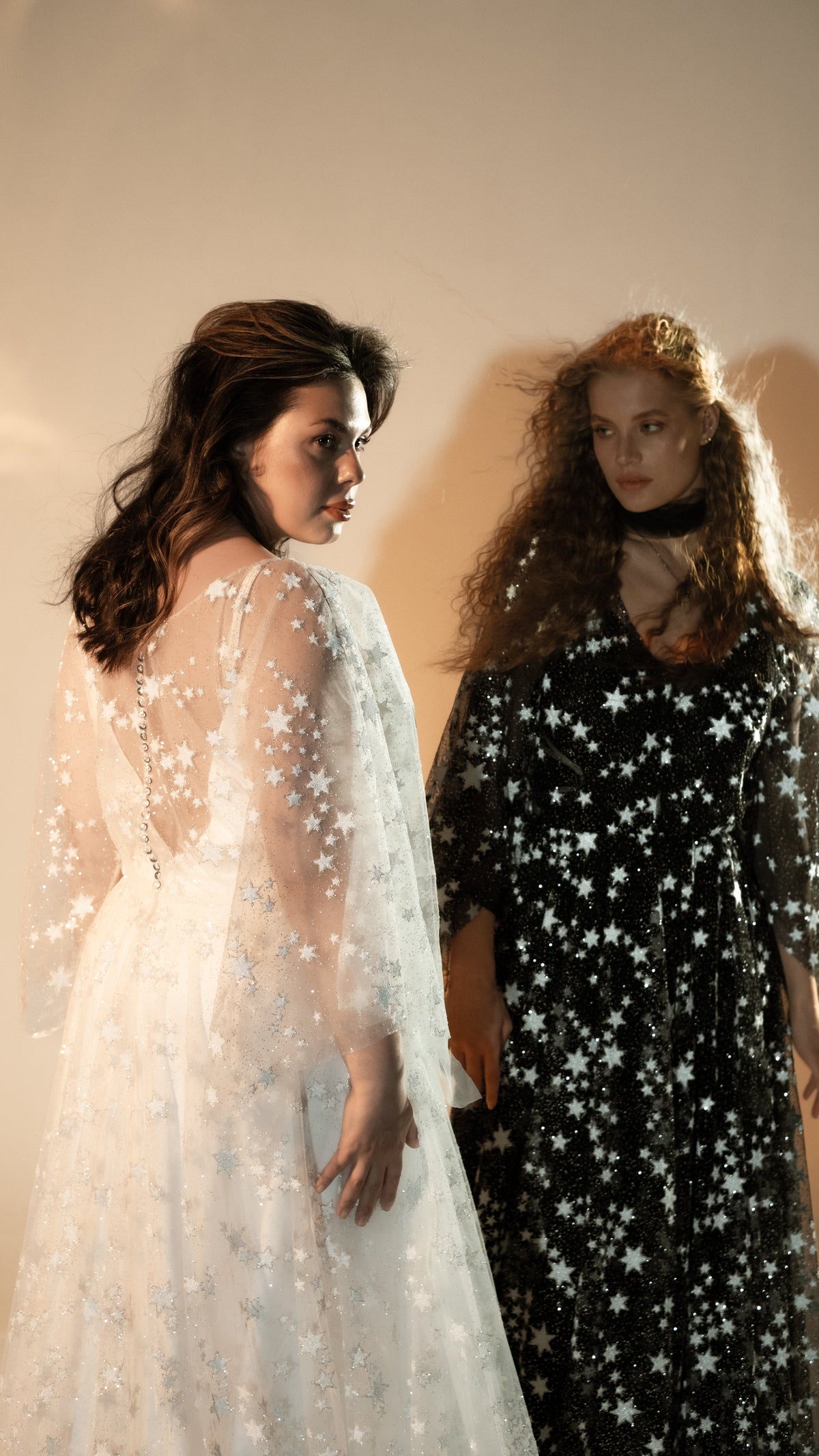 Back view of an ivory star lace wedding dress with buttoned back beside a model in a black star lace gown, Counting Stars 2.0