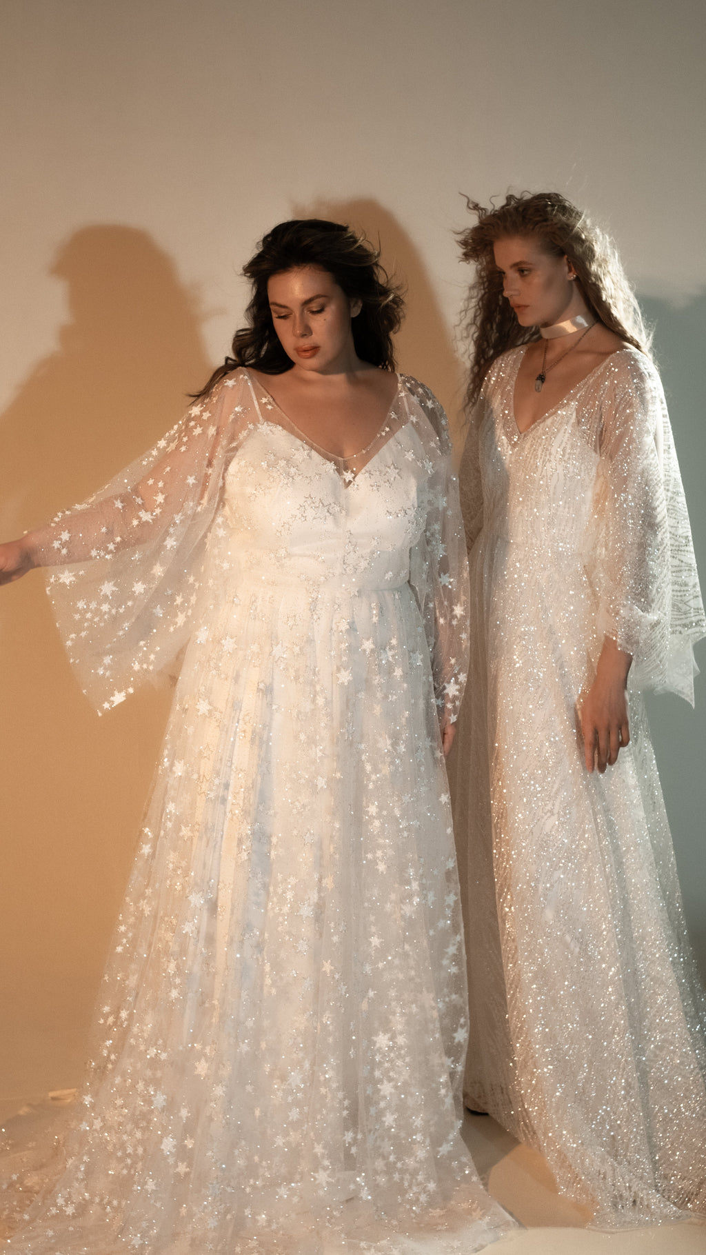 Two brides standing close, one spreading the bell sleeve of a star lace wedding dress, warm studio, Counting Stars 2.0