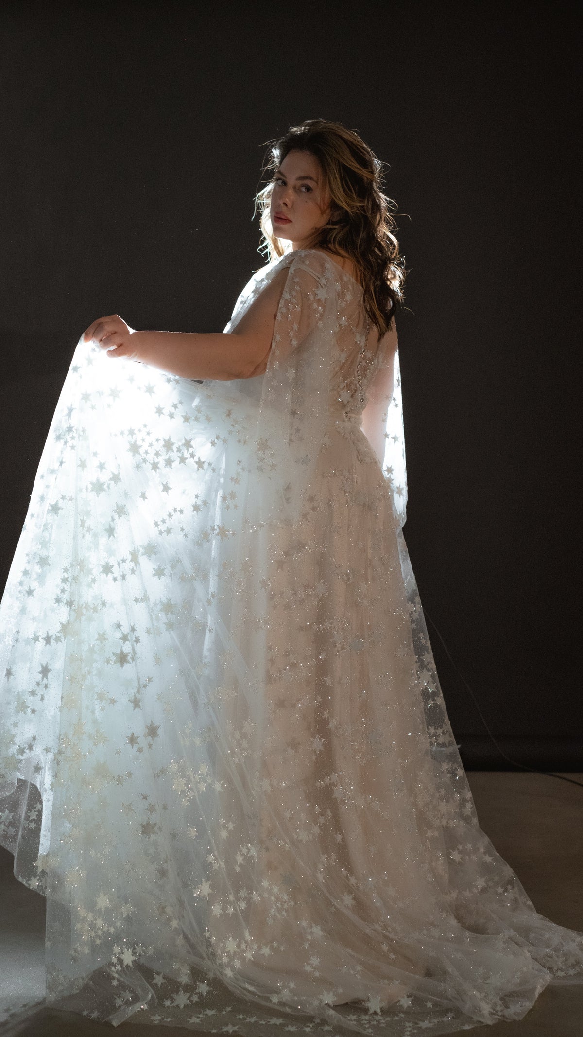 Bride glancing back while lifting the sheer star lace cape sleeve of her wedding dress, dark studio, Counting Stars 2.0