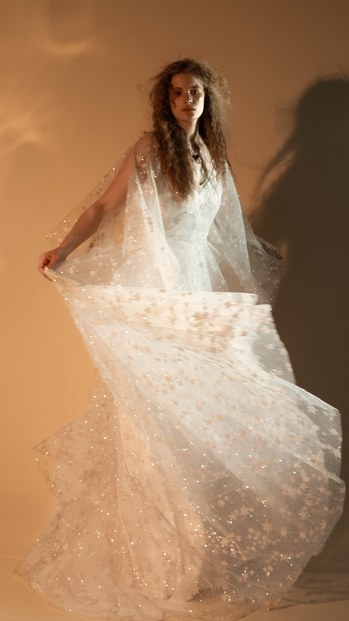 Bride swinging the sheer star lace skirt of her wedding dress out to the side, warm light on a beige wall, Counting Stars 2.0