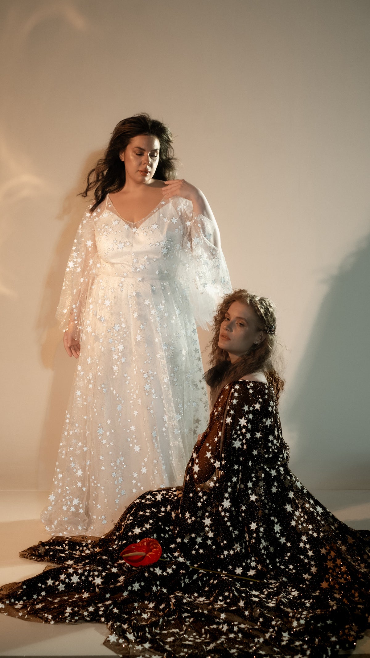 Bride standing in an ivory star lace wedding dress beside a seated model in a black star lace gown, Counting Stars 2.0