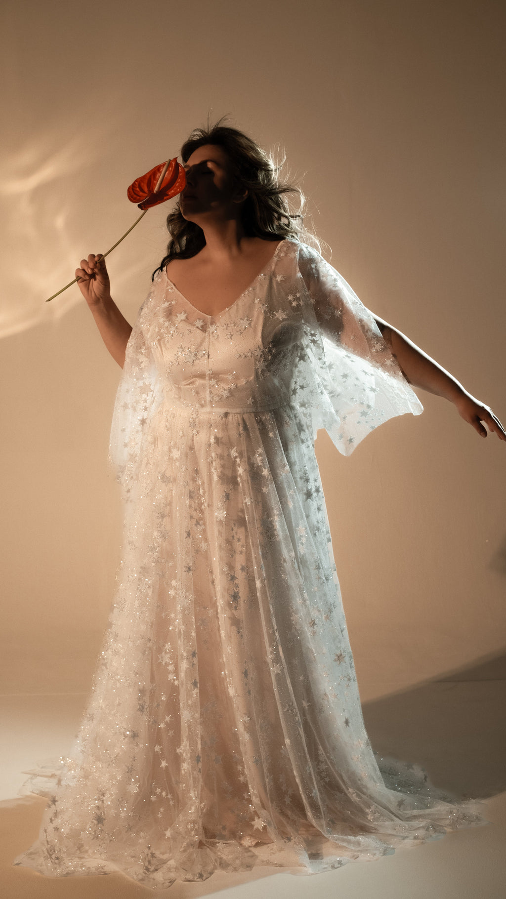 Bride holding a red anthurium to her face, star lace wedding dress with open cape sleeves and a soft train, Counting Stars 2.0