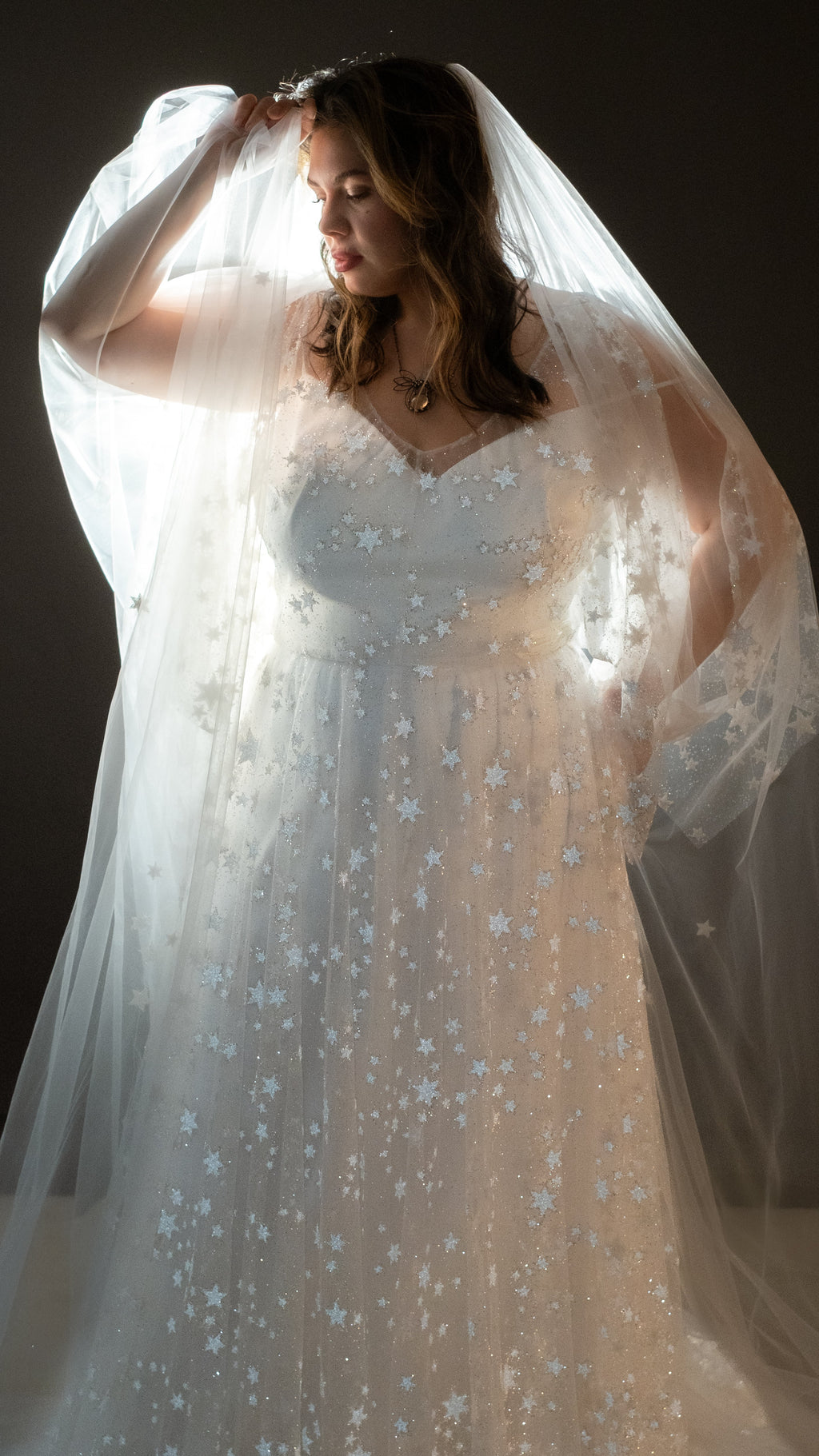 Bride lifting a long tulle veil over her head, backlit star lace wedding dress on a dark backdrop, Counting Stars 2.0