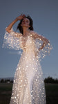 Celestial wedding dress in star lace with sheer bell sleeves, bride posed at dusk with one hand raised. Counting Stars Sample by Ephemeris