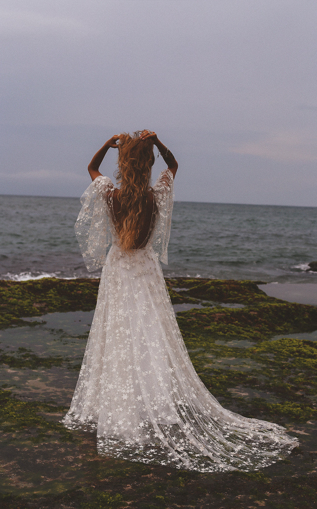 Back view of a bride on a seaweed covered shore, open back star lace wedding dress with a long train, Counting Stars Sample