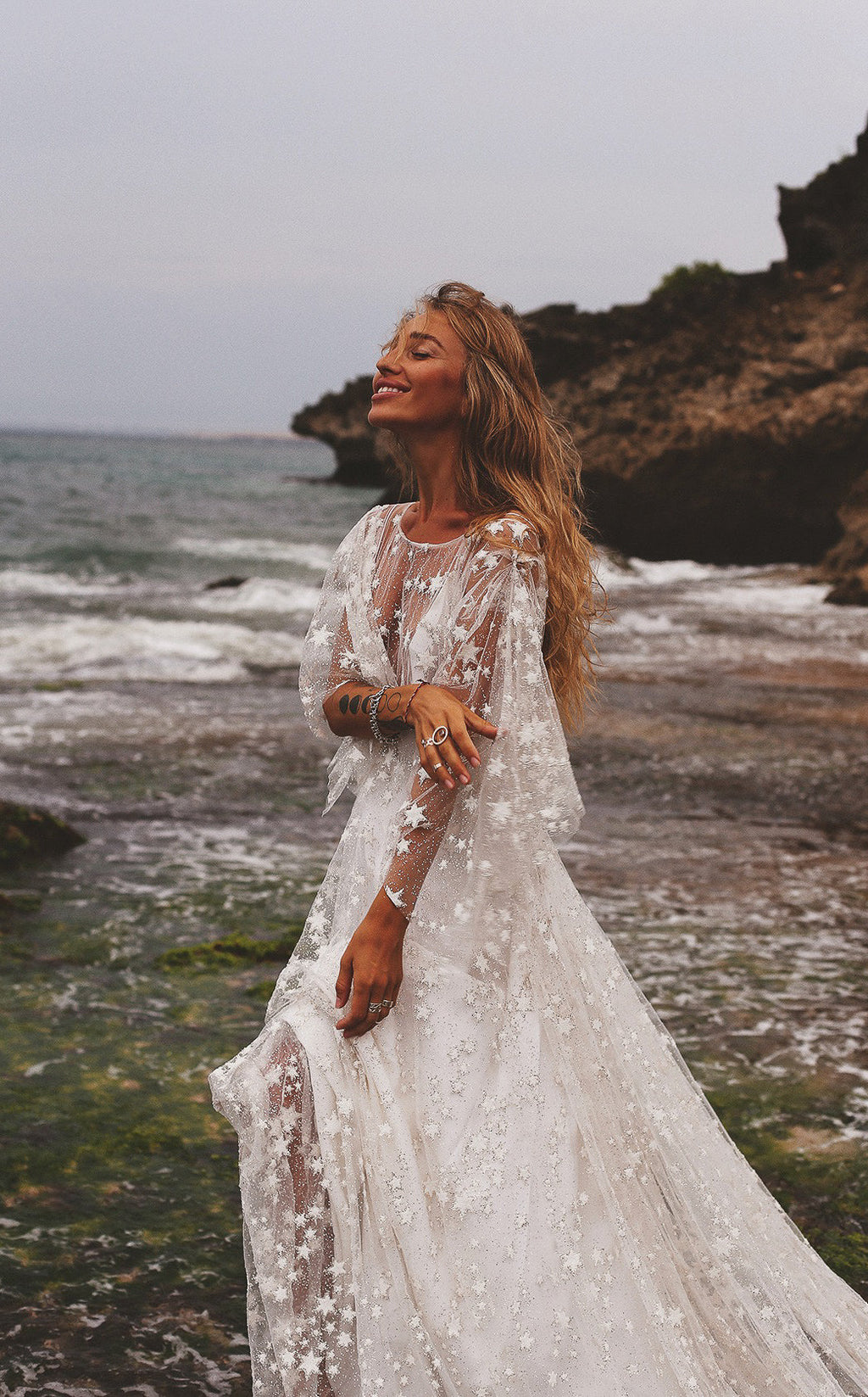 Bride laughing by the sea in a sheer star lace wedding dress with flutter sleeves, cliffs behind, Counting Stars Sample
