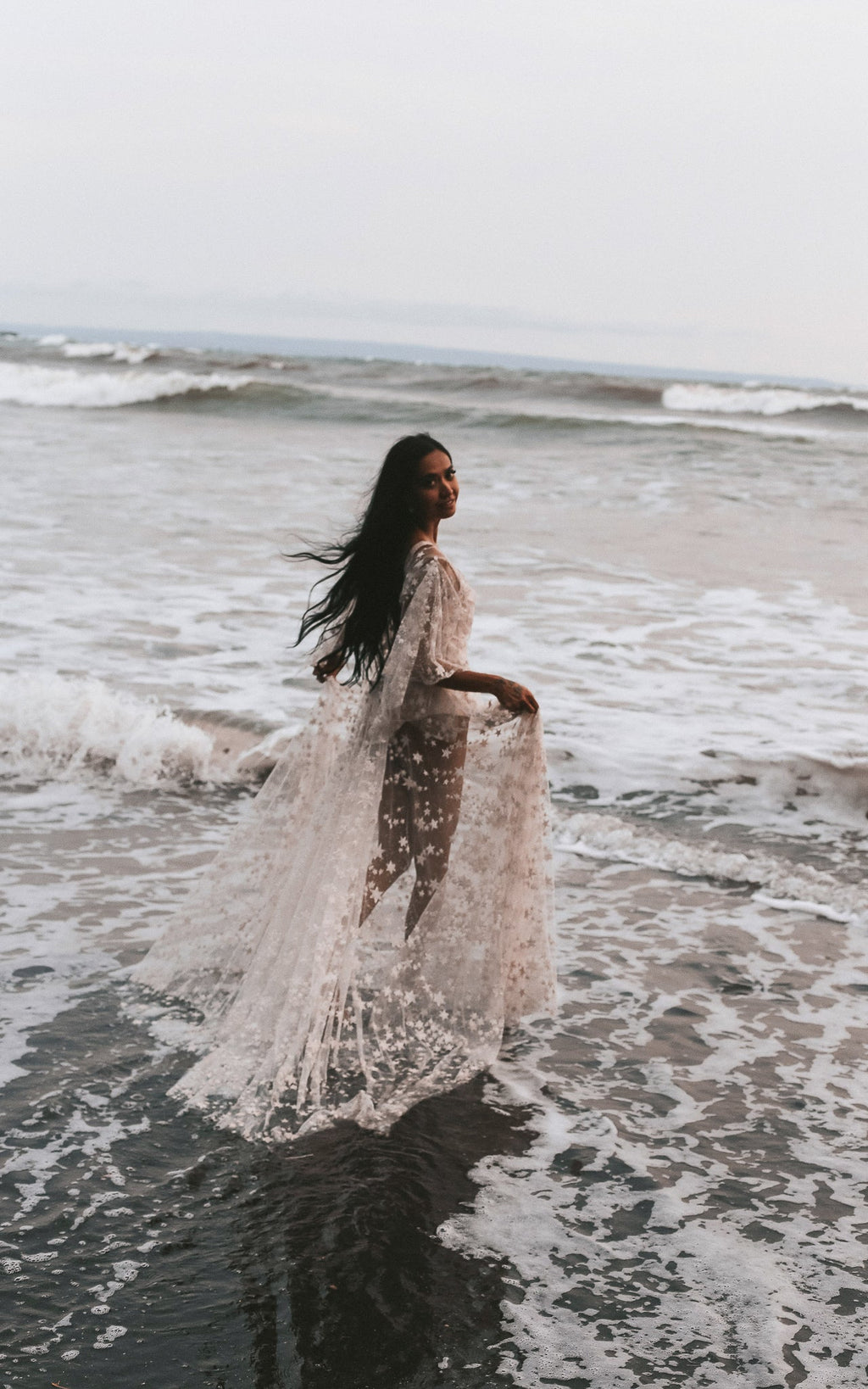 Bride wading through foaming surf, holding up the sheer star lace skirt of her wedding dress, Counting Stars Sample