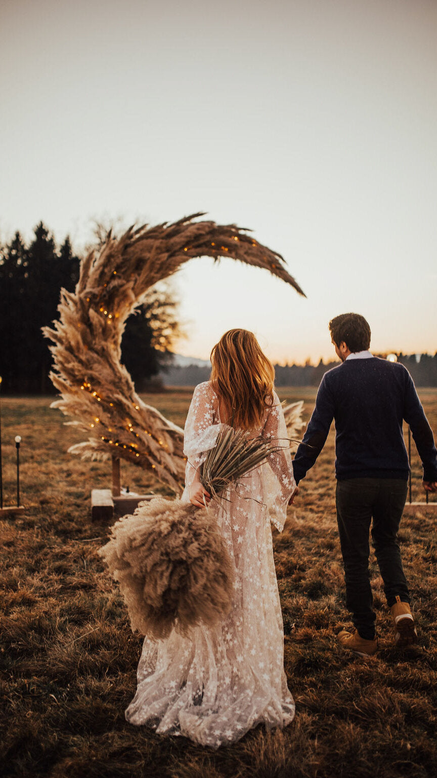 Couple walking toward a pampas grass moon arch at sunset, bride in a star lace wedding dress, Counting Stars Sample