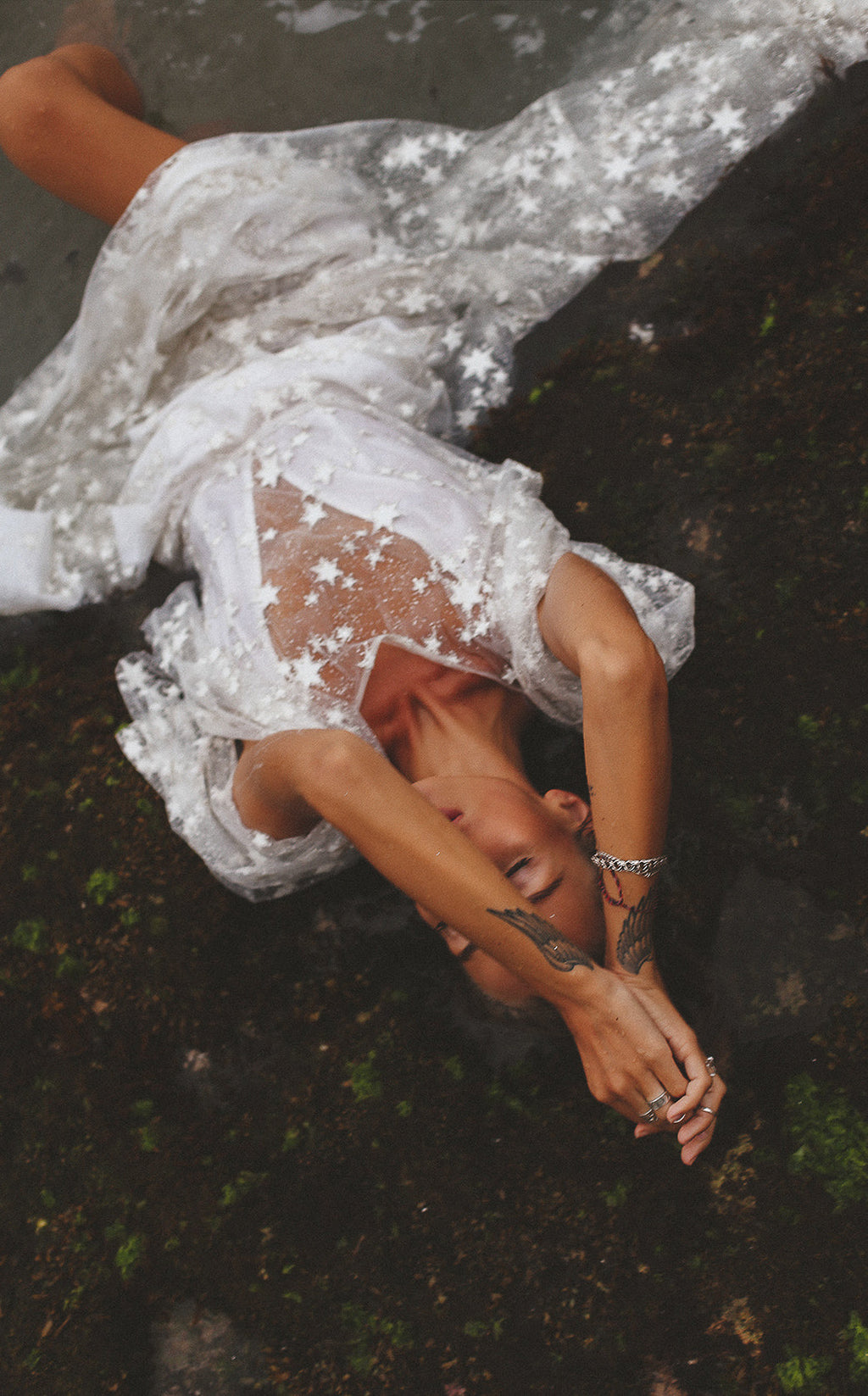 Overhead shot of a bride lying on mossy ground, sheer star lace sleeves of her wedding dress spread out, Counting Stars Sample