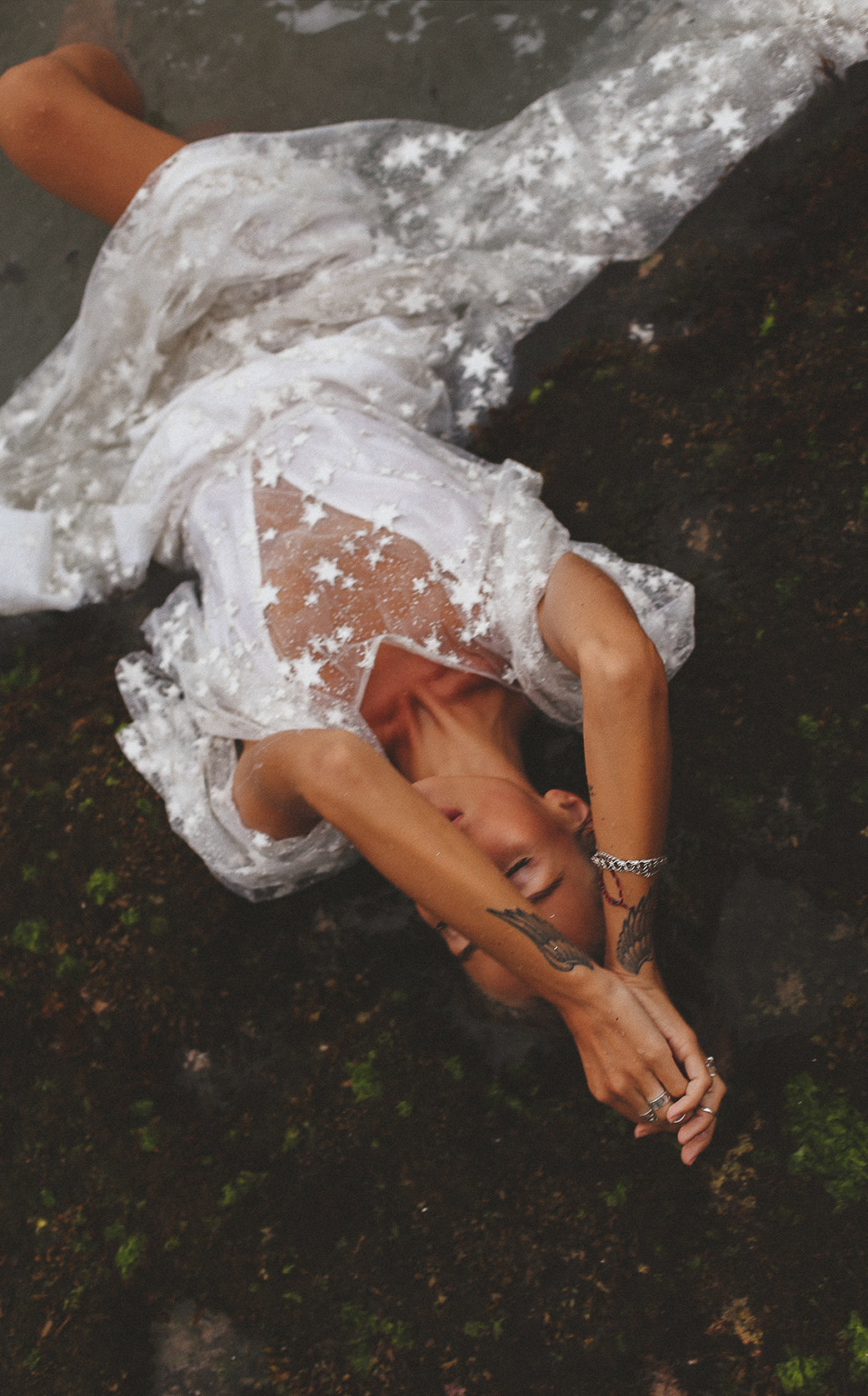 Overhead shot of a bride lying on mossy ground, sheer star lace sleeves of her wedding dress spread out, Counting Stars Sample