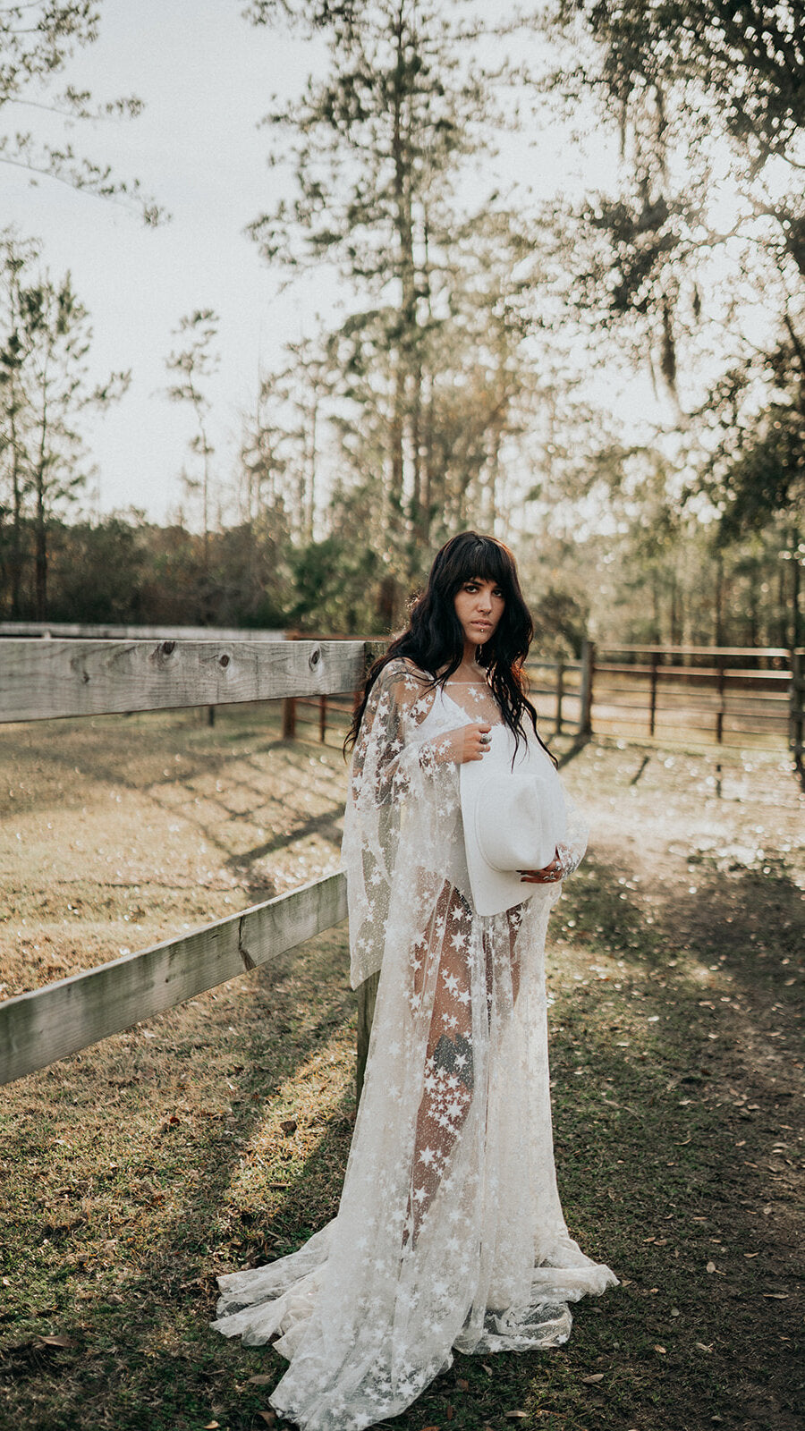 Bride holding a white hat by a ranch fence, sheer star lace wedding dress with bell sleeves and train, Counting Stars Sample