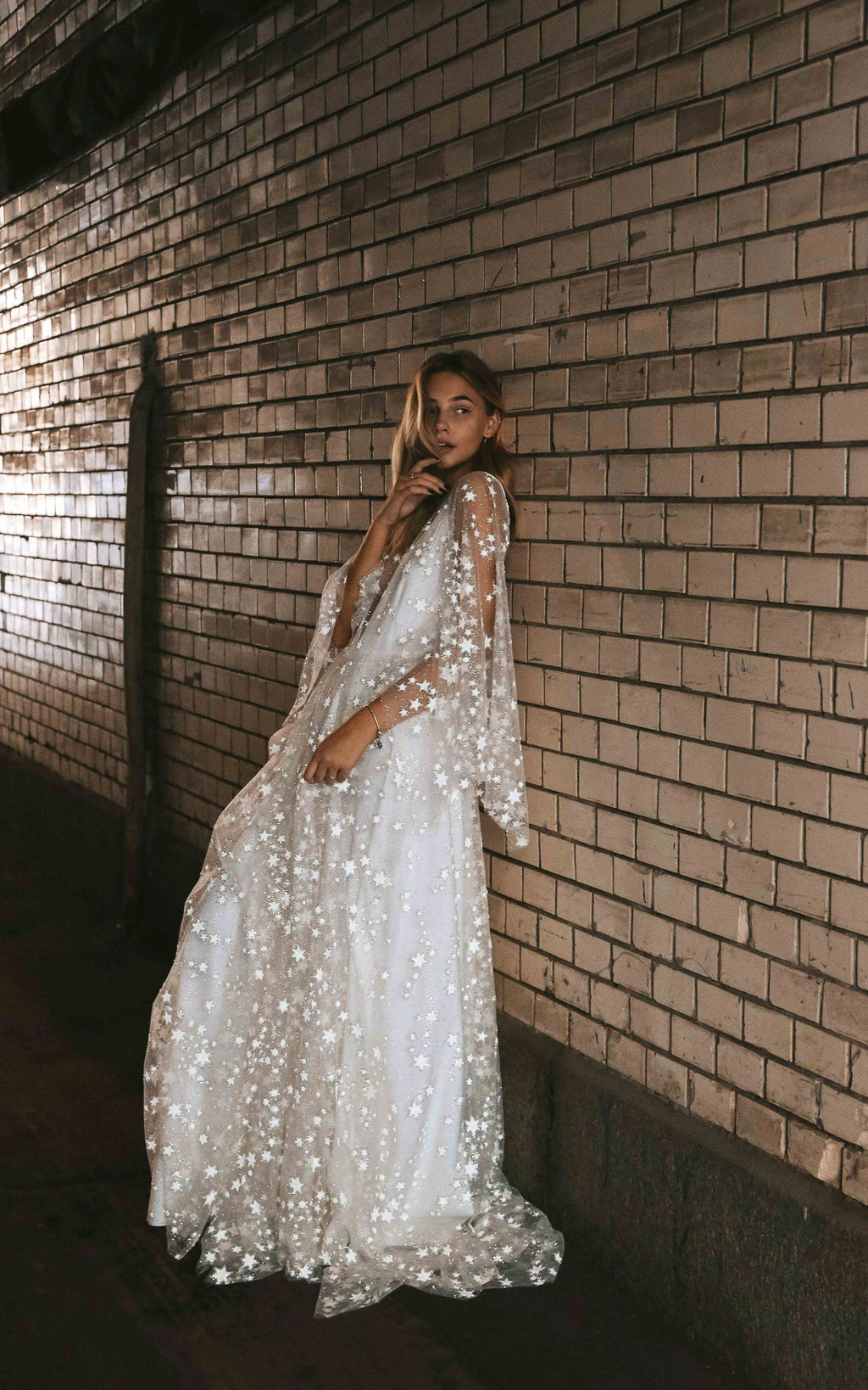 Bride leaning on a brick wall at night in a star lace wedding dress with long cape sleeves and a soft train, Counting Stars