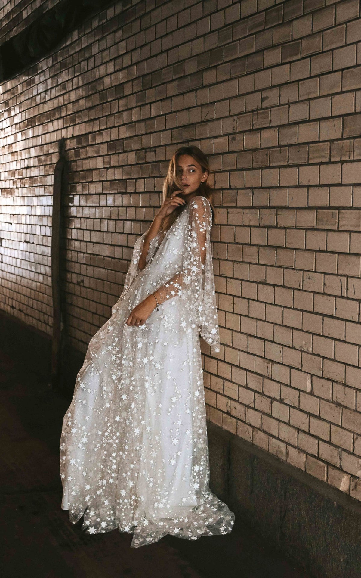 Bride leaning on a brick wall at night in a star lace wedding dress with long cape sleeves and a soft train, Counting Stars