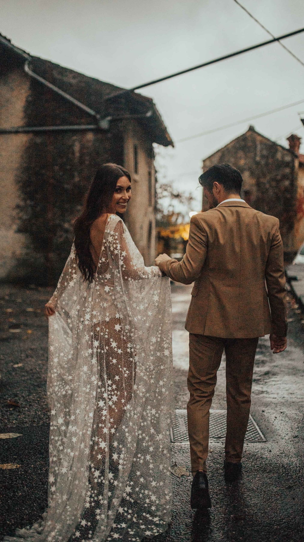 Bride looking back with a smile, open back star lace wedding dress with long sleeves on a village street, Counting Stars