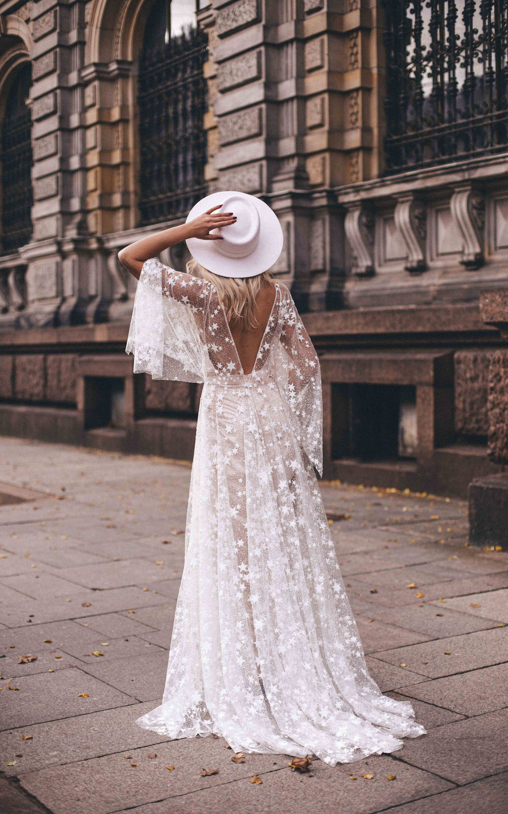 Back view of a bride in a white hat on a city pavement, open V-back star lace wedding dress with flutter sleeves, Counting Stars
