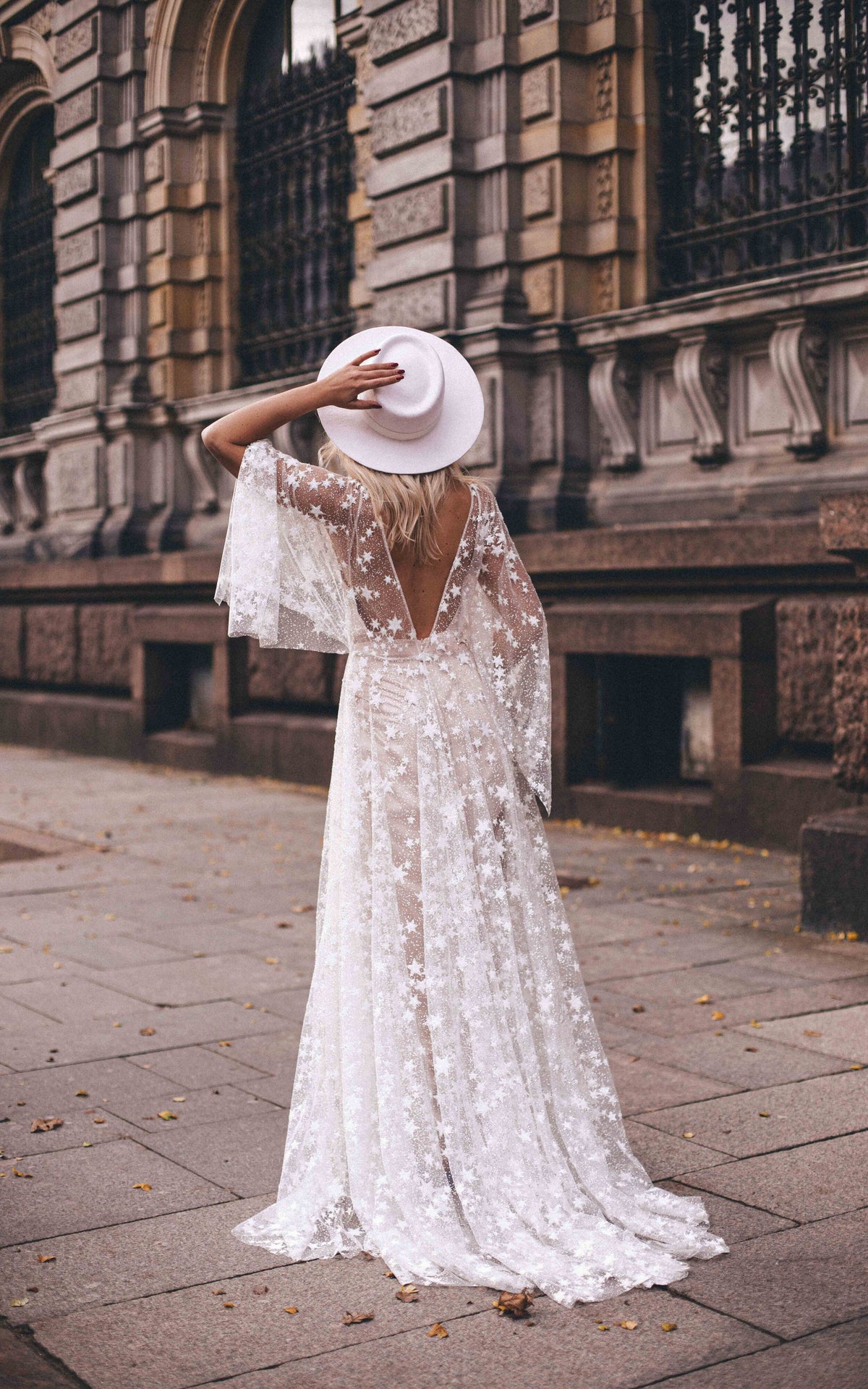 Back view of a bride in a white hat on a city pavement, open V-back star lace wedding dress with flutter sleeves, Counting Stars