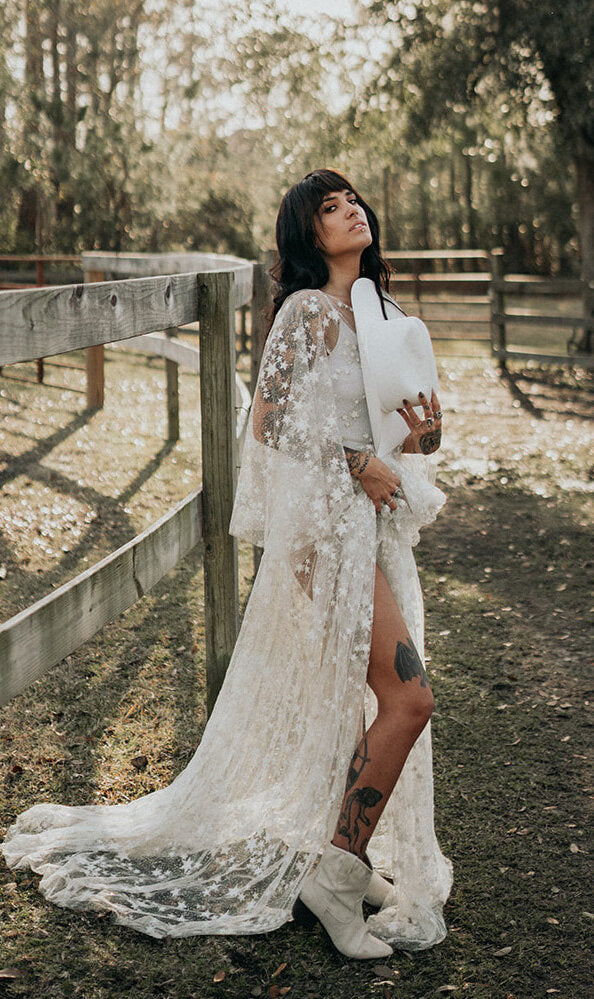 Bride by a wooden ranch fence in a star lace wedding dress with a leg slit, white boots and a white hat in hand, Counting Stars