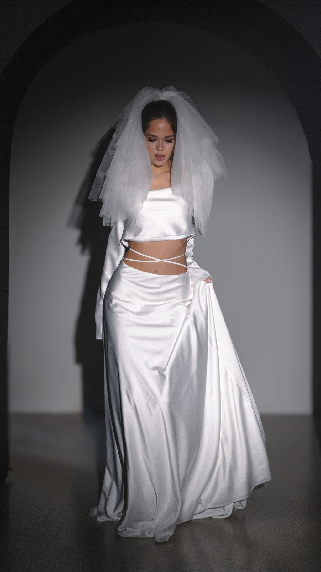 Two-piece silk satin wedding dress, long sleeve crop top and bias skirt with crossed waist ties, short tulle veil, studio arch. Danielle by Ephemeris