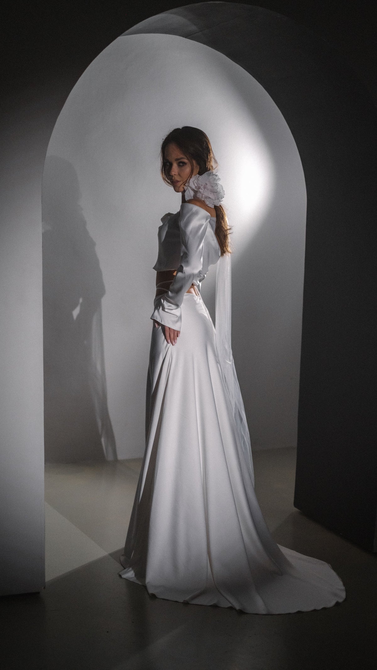 Back view of an ivory silk satin wedding dress with a long sleeve crop top, floor-length skirt and train in an arched doorway, Danielle