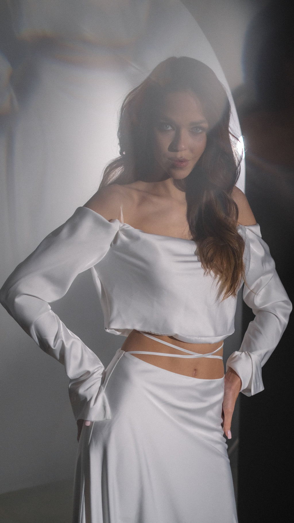 Off-shoulder silk satin crop top with long sleeves and a satin bridal skirt with waist ties, hand on hip, Danielle