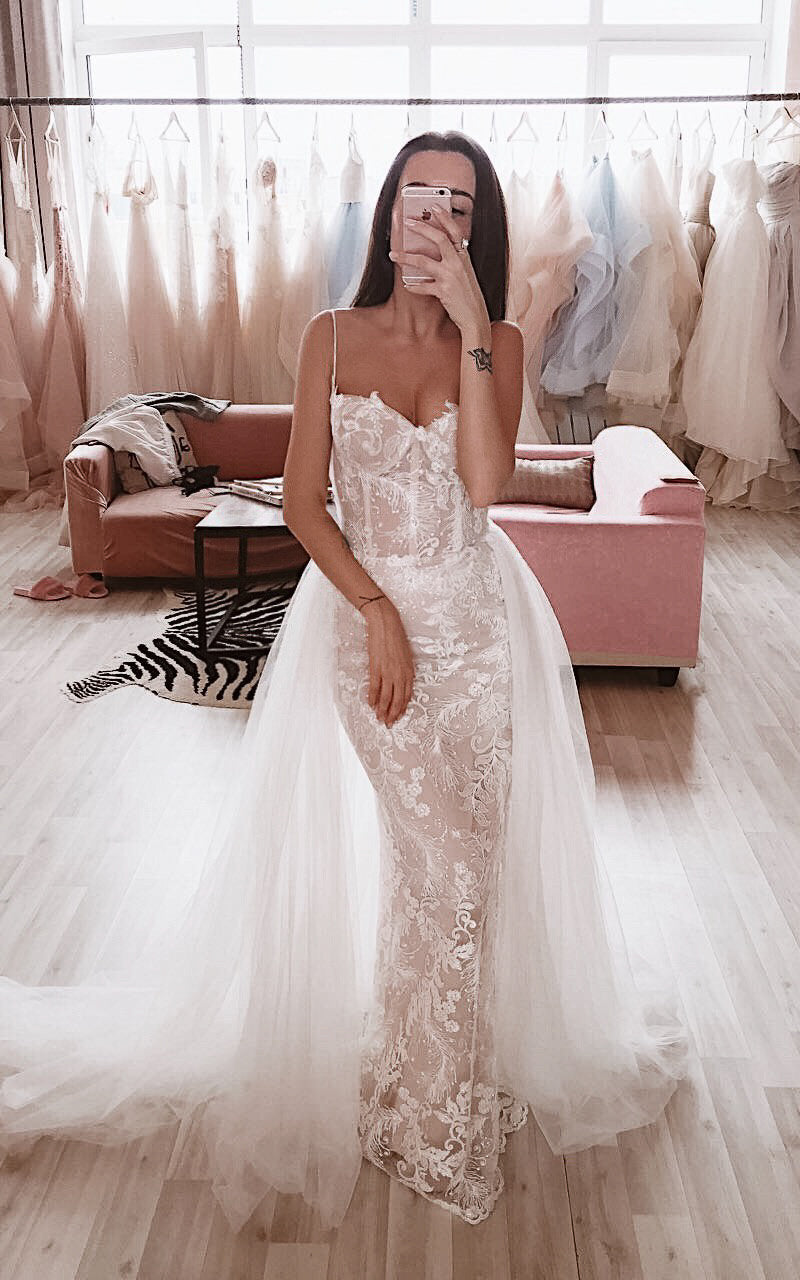 Lace mermaid wedding dress with thin straps, beige lining and a full tulle overskirt, mirror selfie in a bridal studio. Feathers Sample by Ephemeris