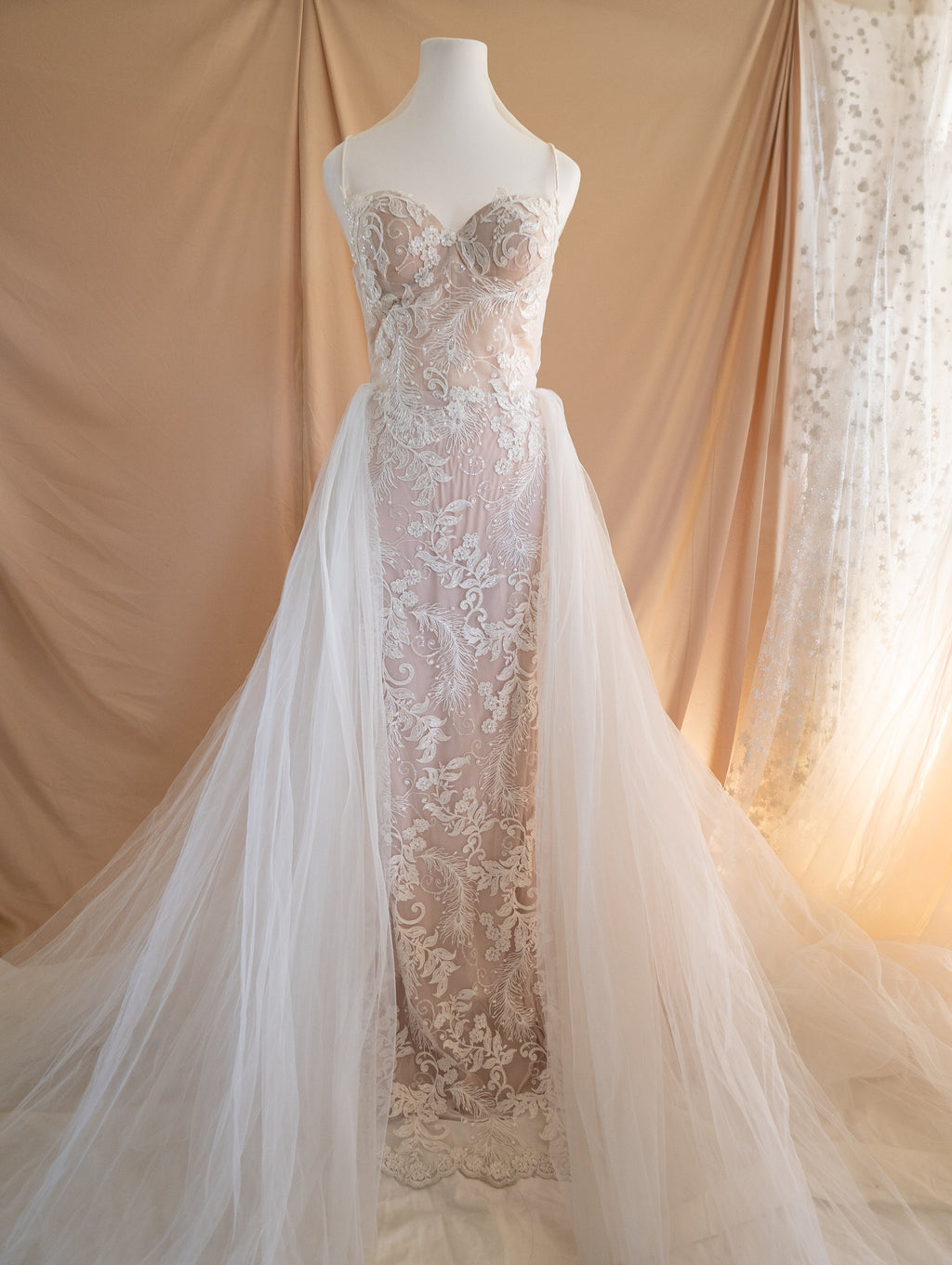 Full-length lace wedding dress on a dress form with a long tulle overskirt, beige drape backdrop, Feathers Sample