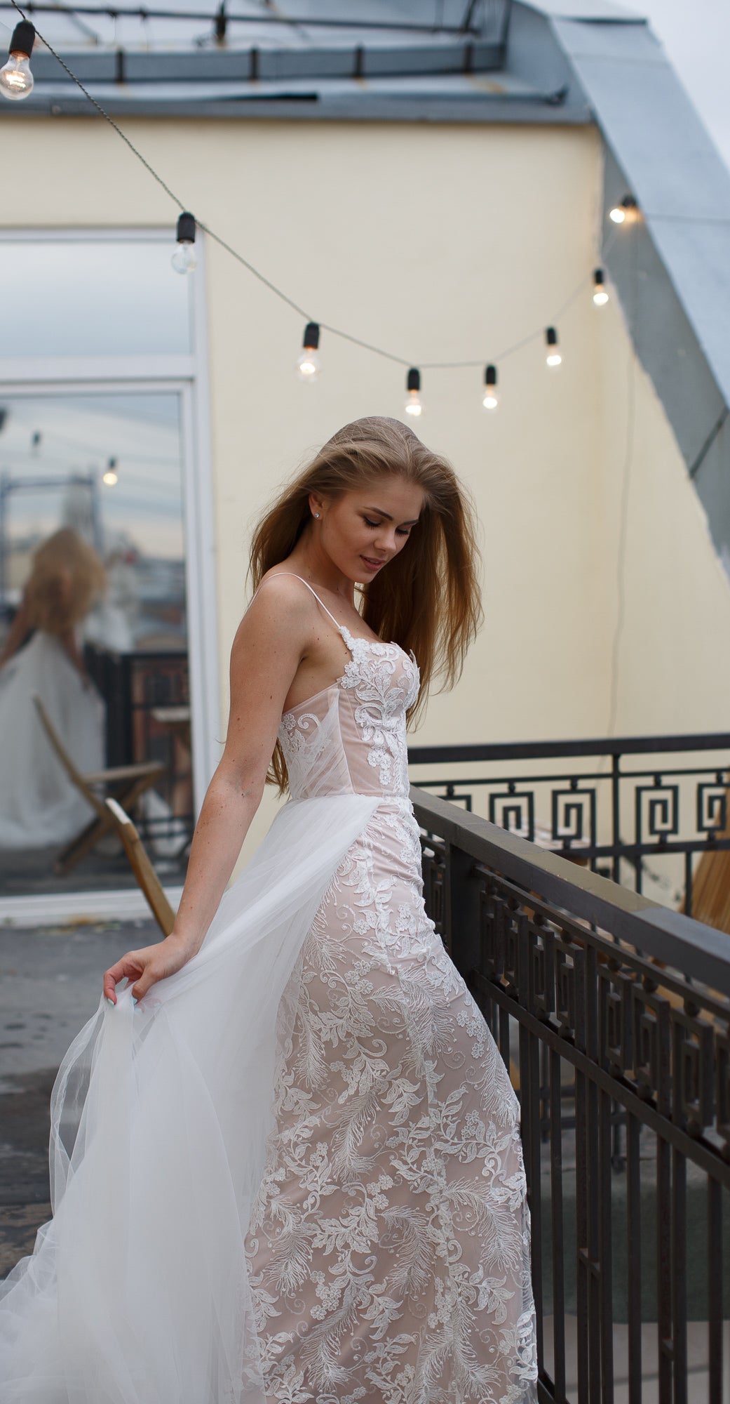 Bride looking down and holding out the tulle overskirt of a lace mermaid wedding dress, terrace lights, Feathers Sample