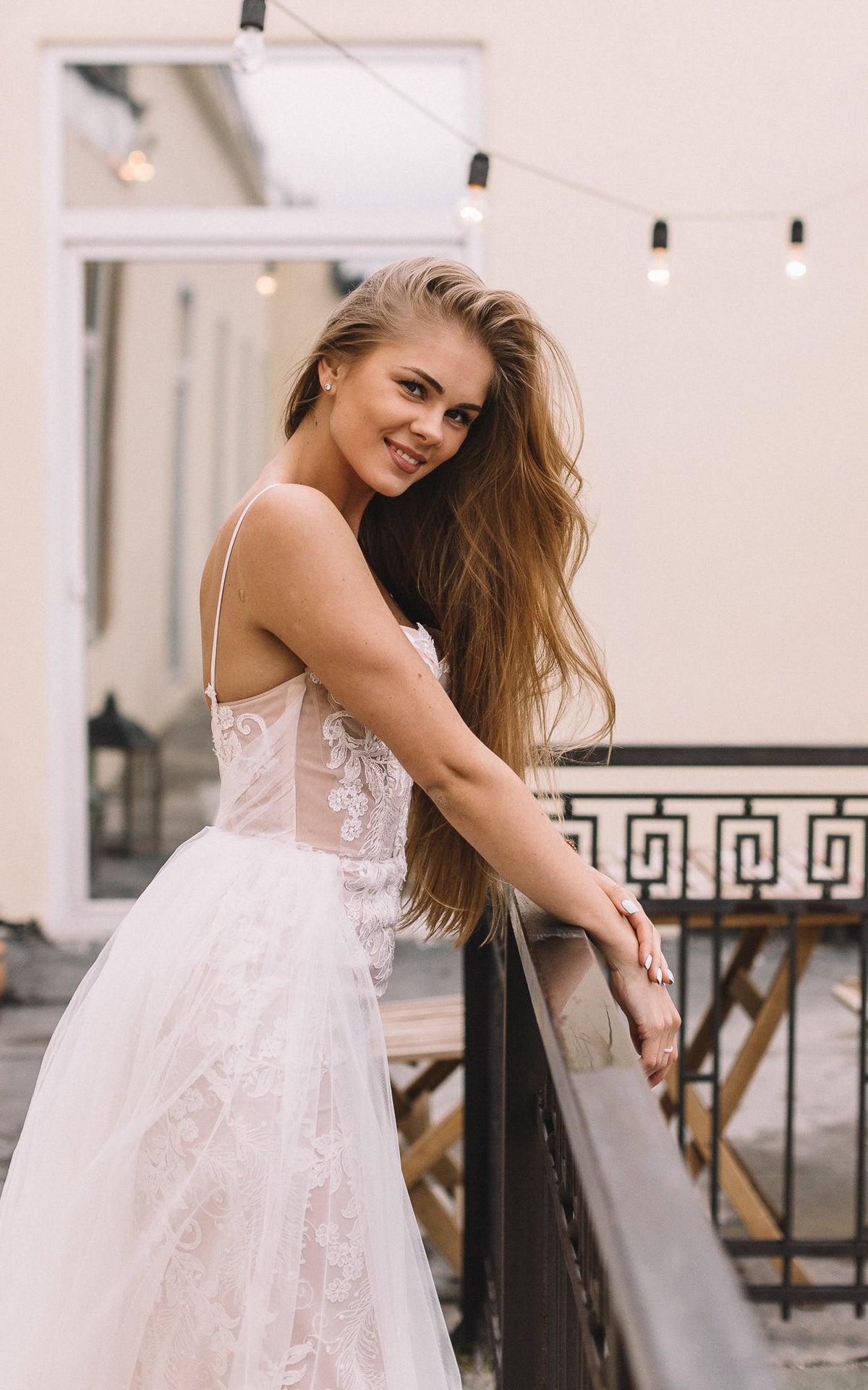 Bride resting both hands on a railing, lace wedding dress with spaghetti straps and soft tulle overlay, Feathers Sample