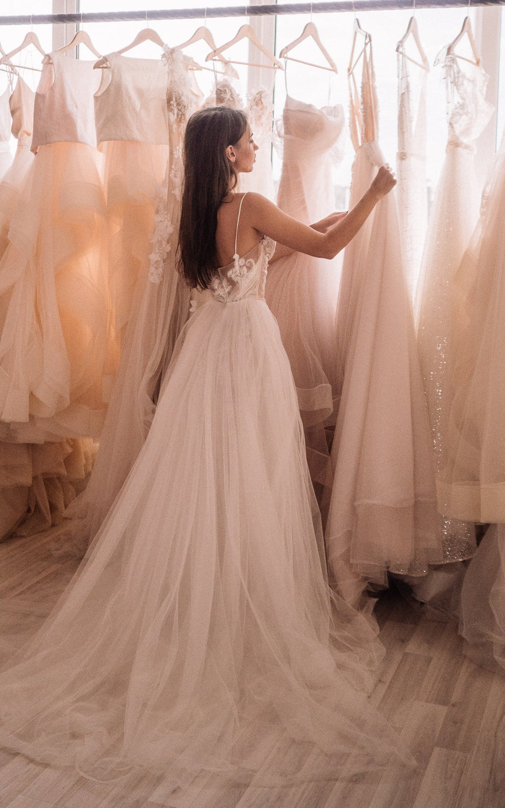 Back view of a bride touching gowns on a rail, strappy back of a wedding dress and a tulle train, Feathers Sample