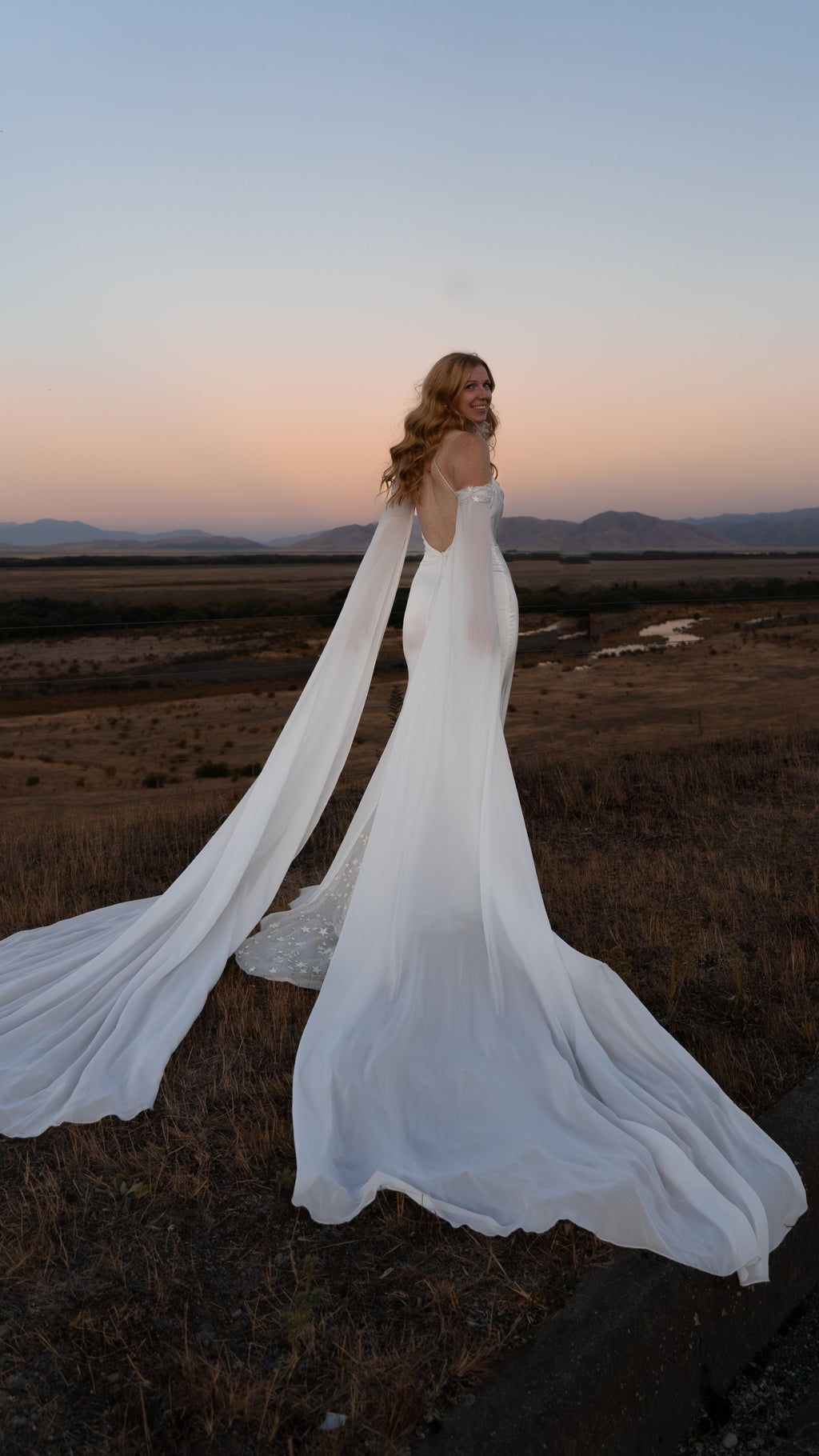 Bride glancing over her shoulder at dusk, silk wedding dress with off-shoulder chiffon sleeves fanned behind, FEYRE