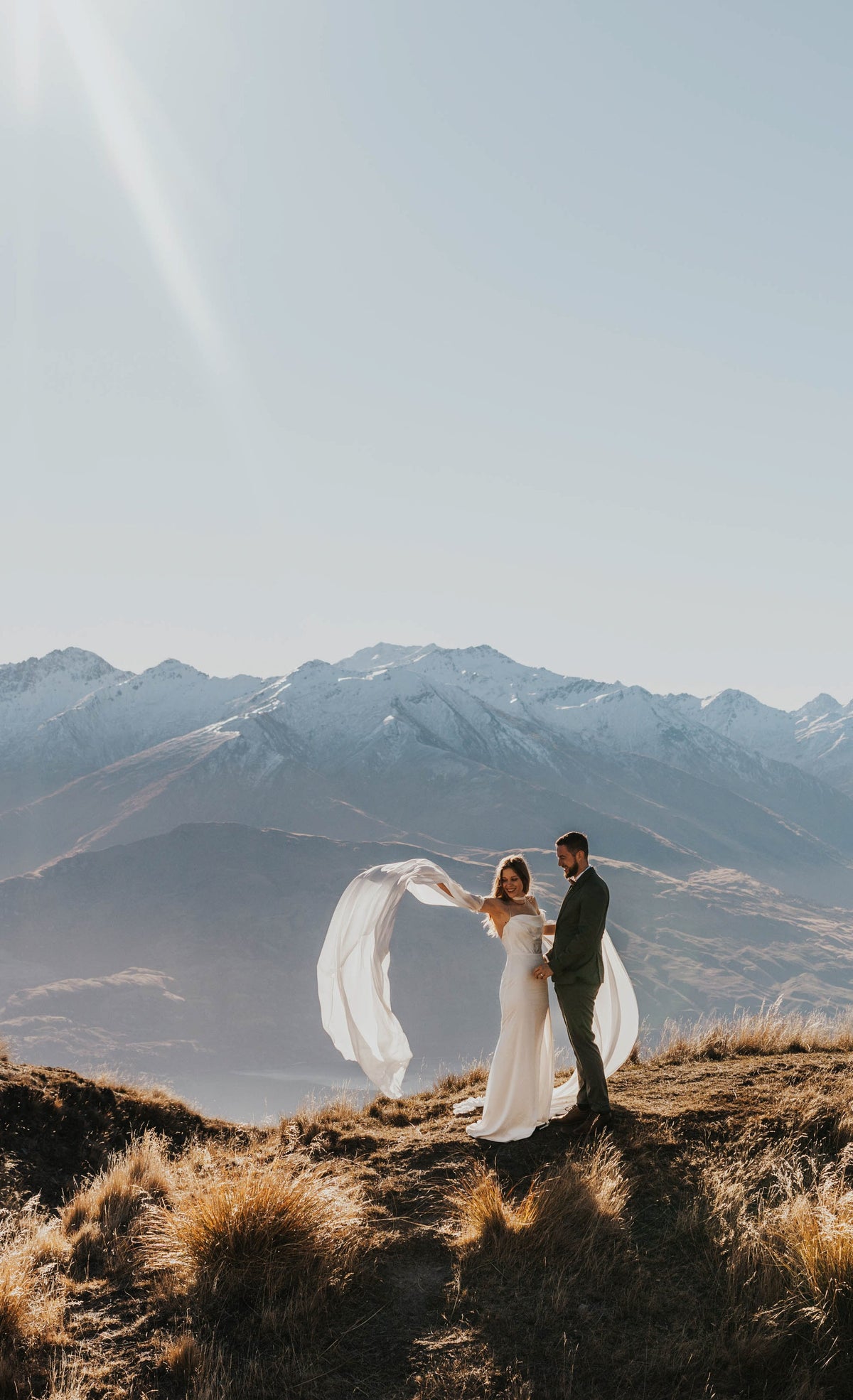 Elopement couple on a mountain top, bride lifting a long chiffon sleeve of her silk wedding dress in the wind, FEYRE