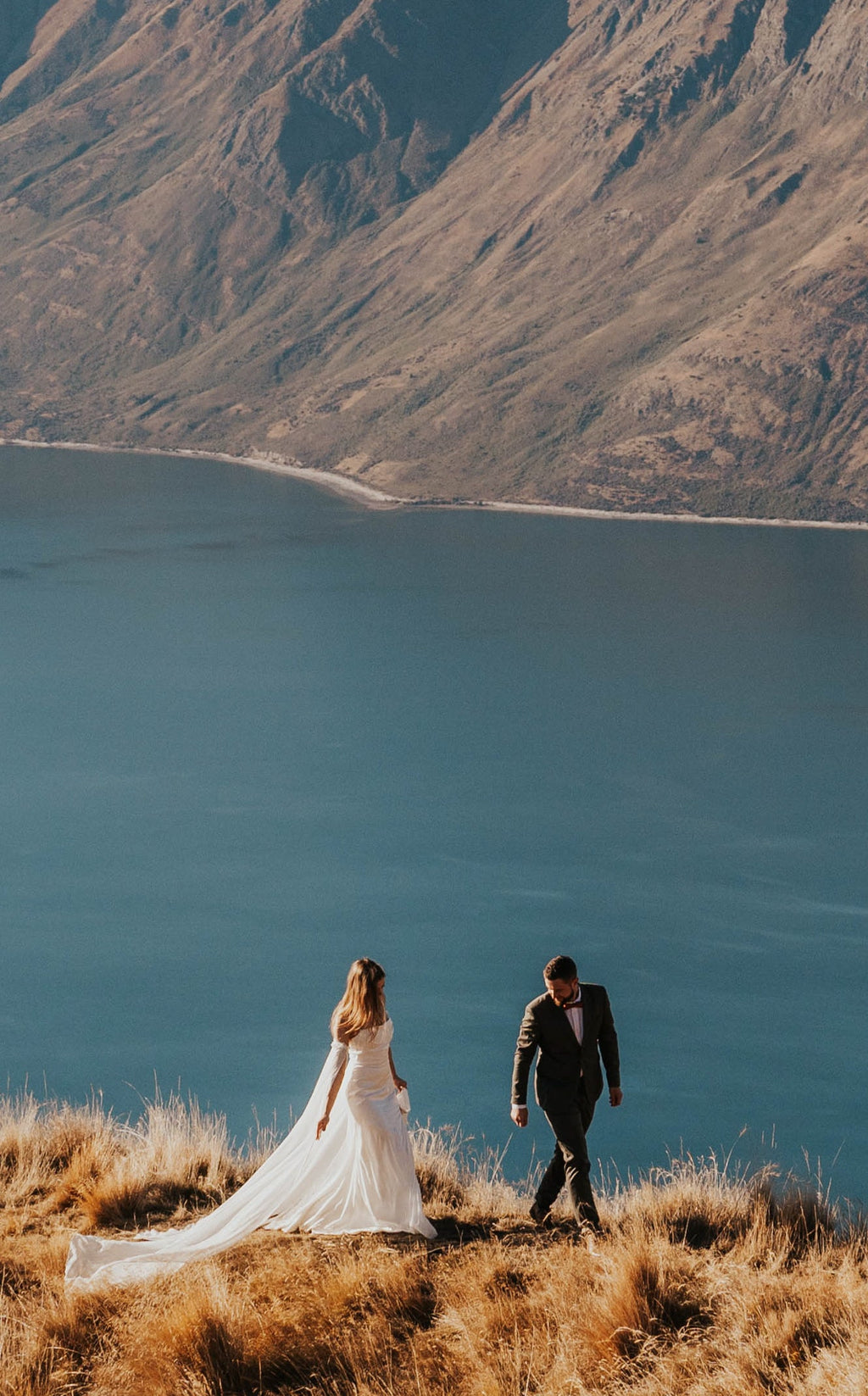 Couple walking along a grassy ridge above a blue lake, bride in a silk wedding dress with trailing sleeves, FEYRE